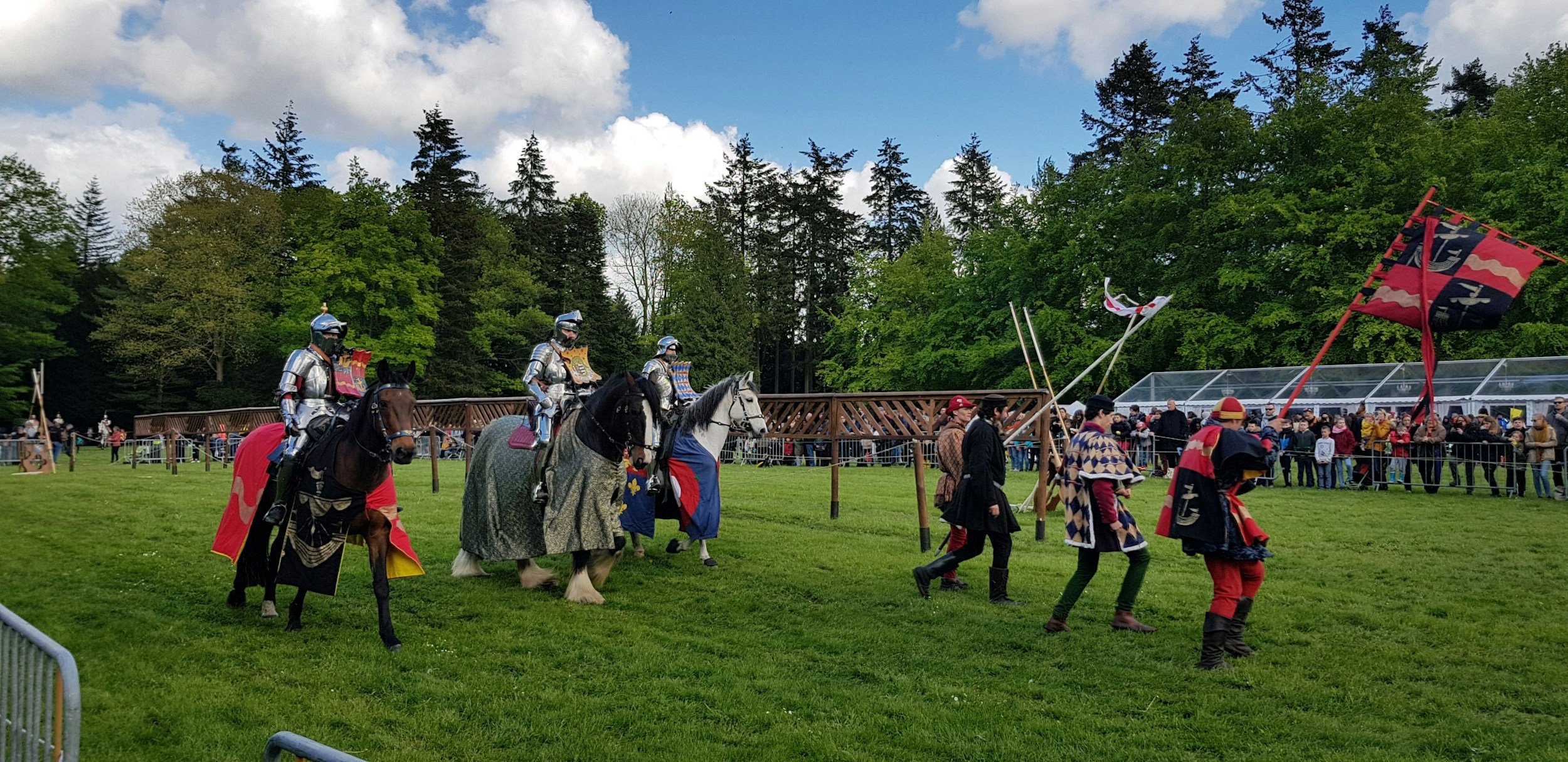 Medieval reenactors dressed in armor and costumes leading horses, with a crowd watching behind a fence at an outdoor event under a partly cloudy sky.