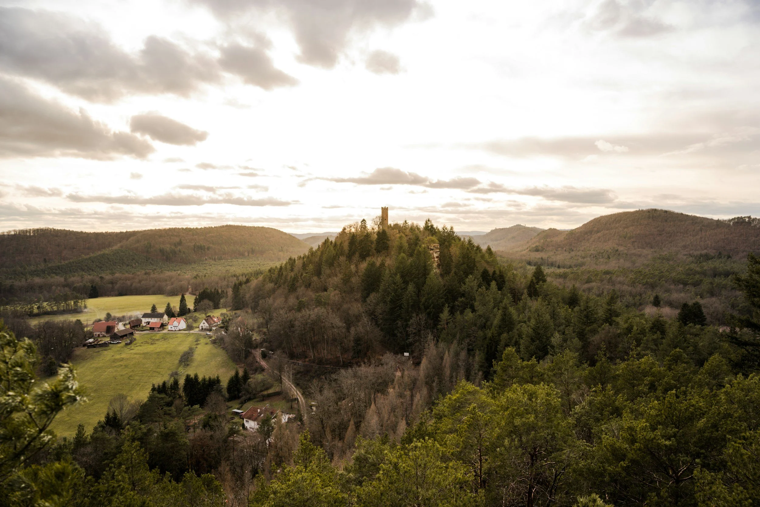 Landscape of hilly countryside with a castle tower on top of a hill and small houses at the base, under a cloudy sky.