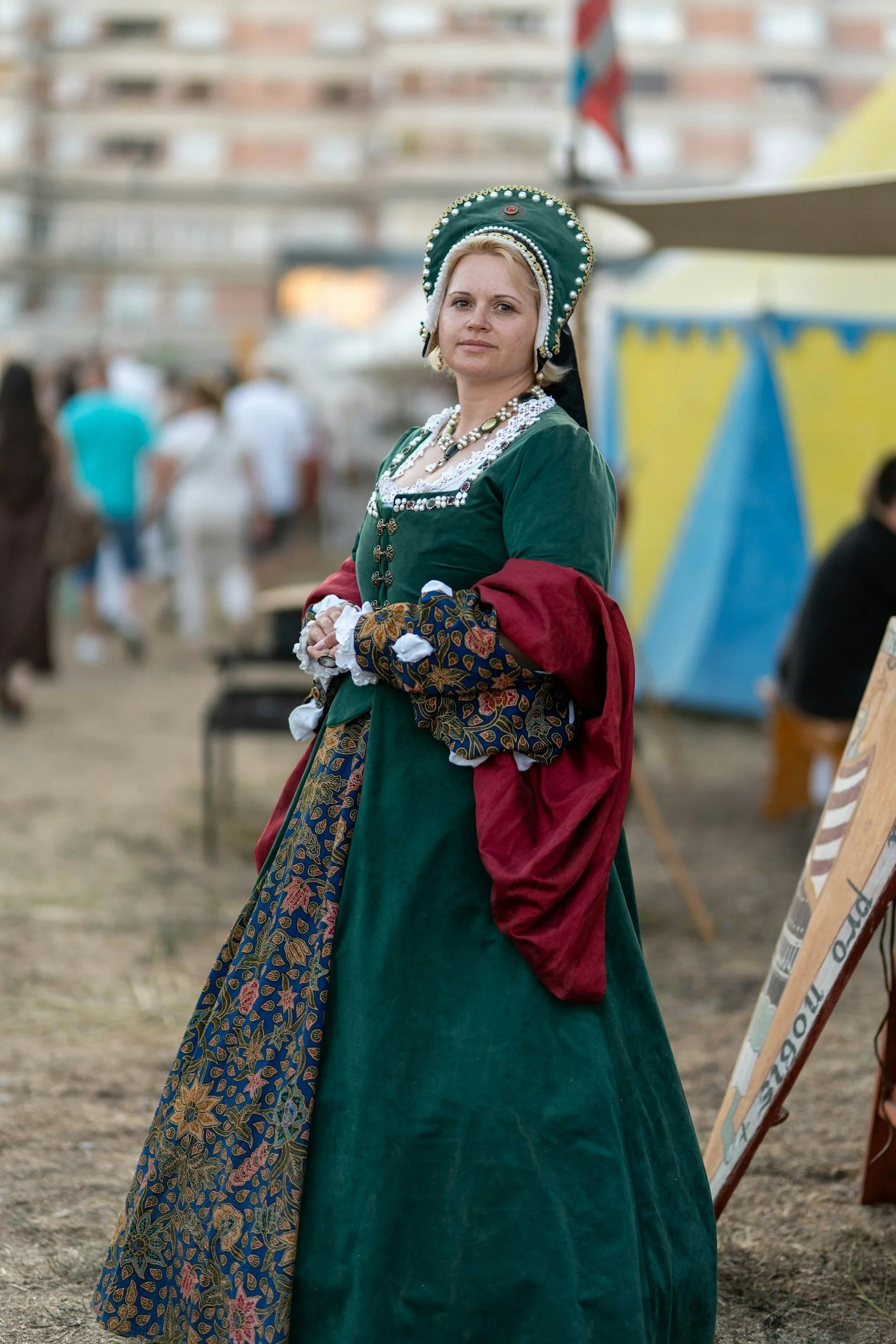 Woman dressed in a traditional green and patterned dress with a matching headpiece and jewelry, standing outdoors at a festival or cultural event.