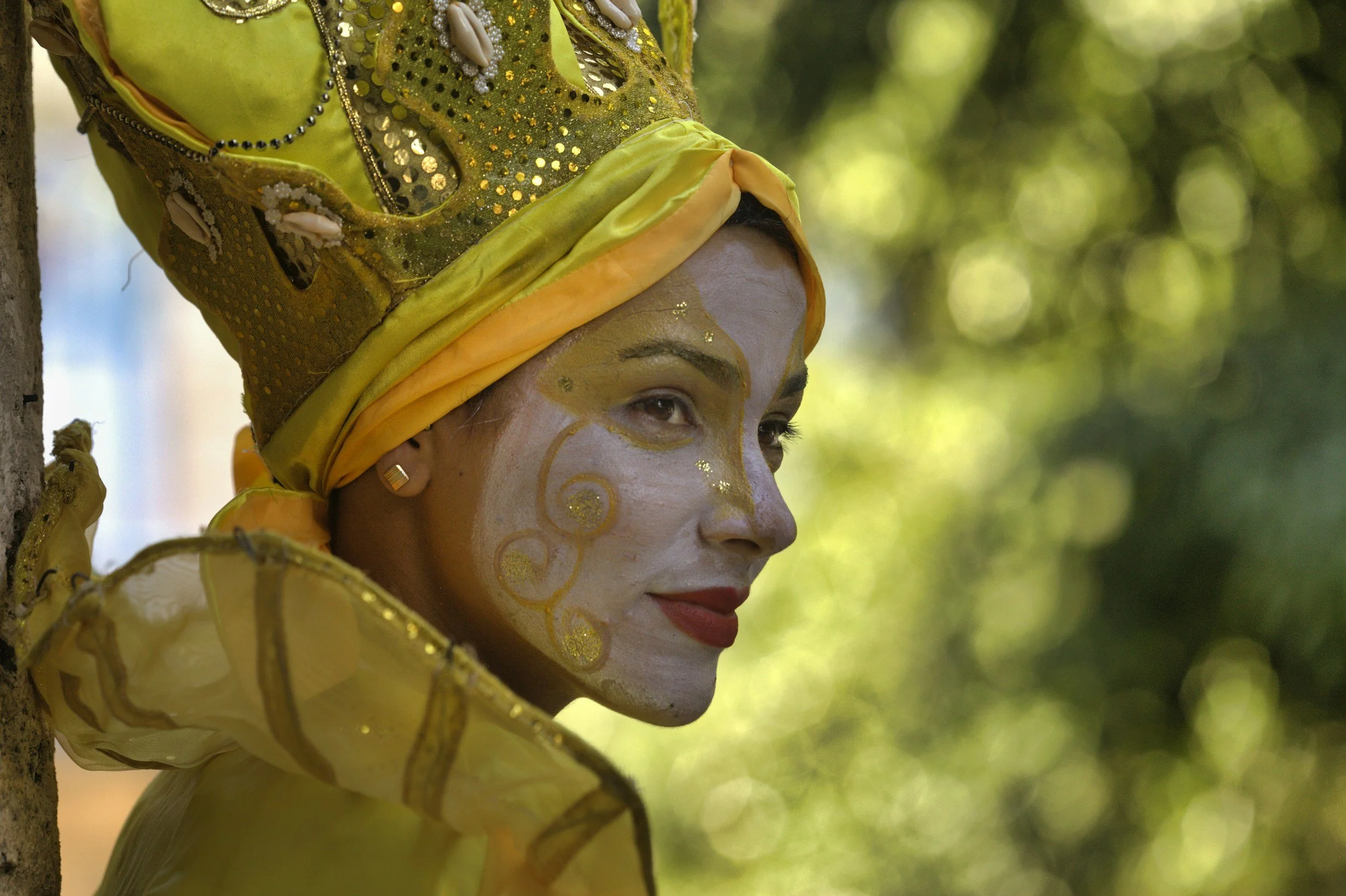 A person wearing vibrant, traditional yellow attire and a headdress, with facial makeup and face paint, outdoors with a blurred green background.