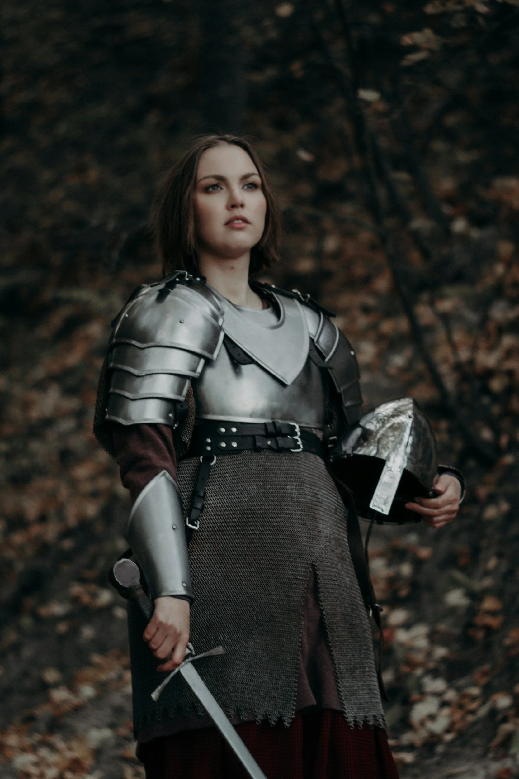 A woman in medieval armor holding a shield and sword in a wooded area with fallen leaves.