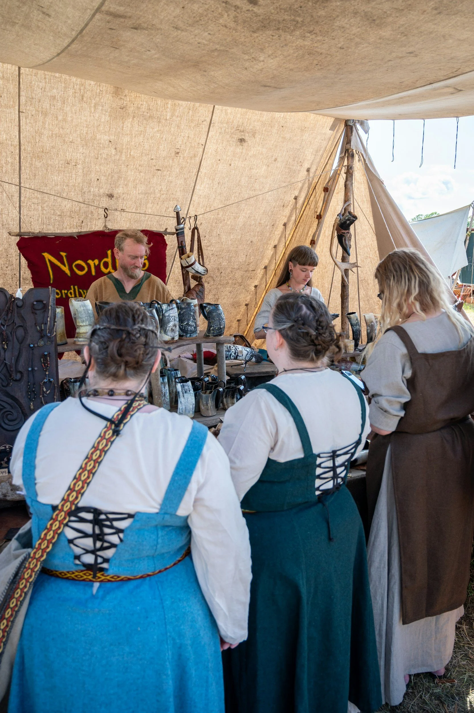 People dressed in medieval or Viking-style clothing inside a tent, browsing and purchasing jewelry and artifacts at a marketplace or fair.