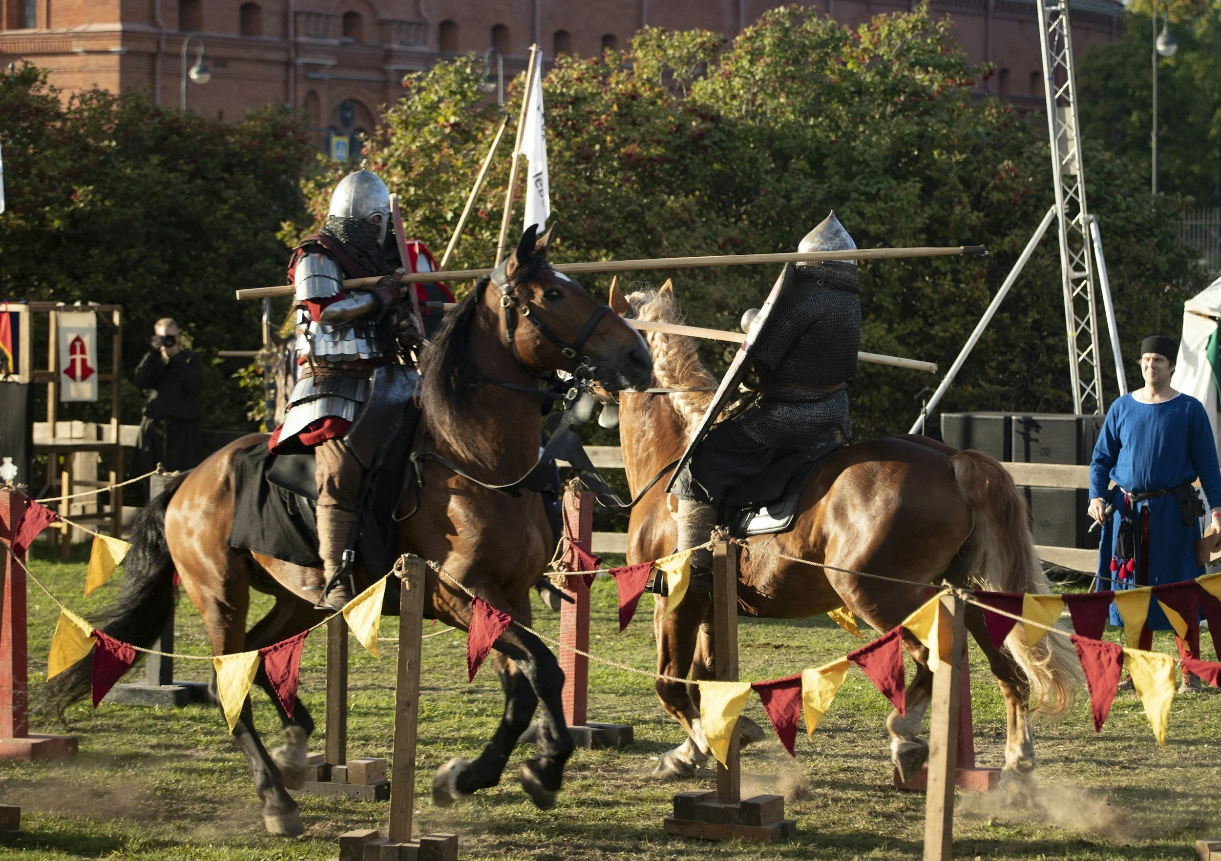 Two knights in armor riding horses during a medieval reenactment, with spectators and a castle in the background.