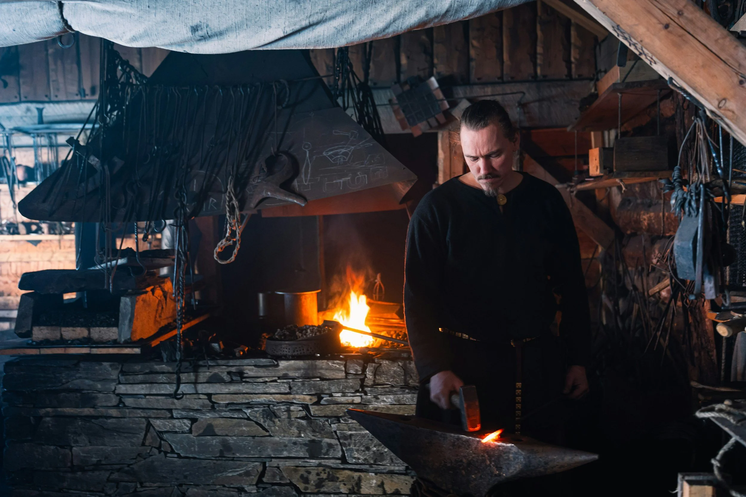 A blacksmith working in a rustic workshop with a fire in the forge, surrounded by tools and equipment, under a wooden roof.