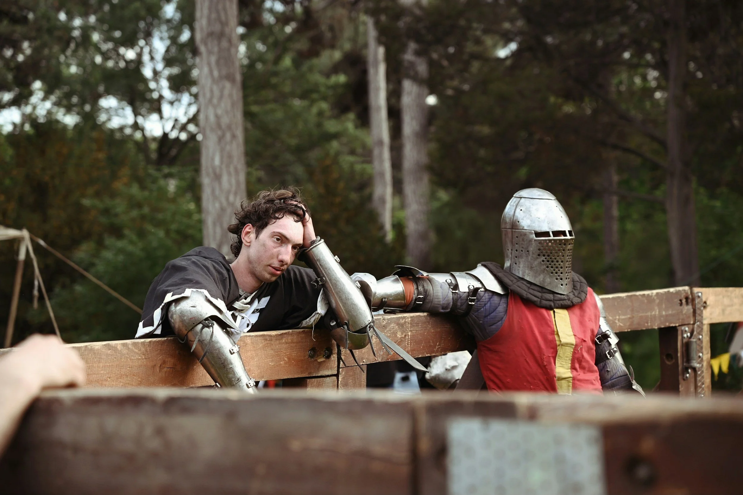 A young man with short curly hair resting his head on his hand at a medieval martial arts event, dressed in a black and white outfit with metal armor on his arms, leaning on a wooden barrier. An armored figure in a metal helmet and red and yellow clo