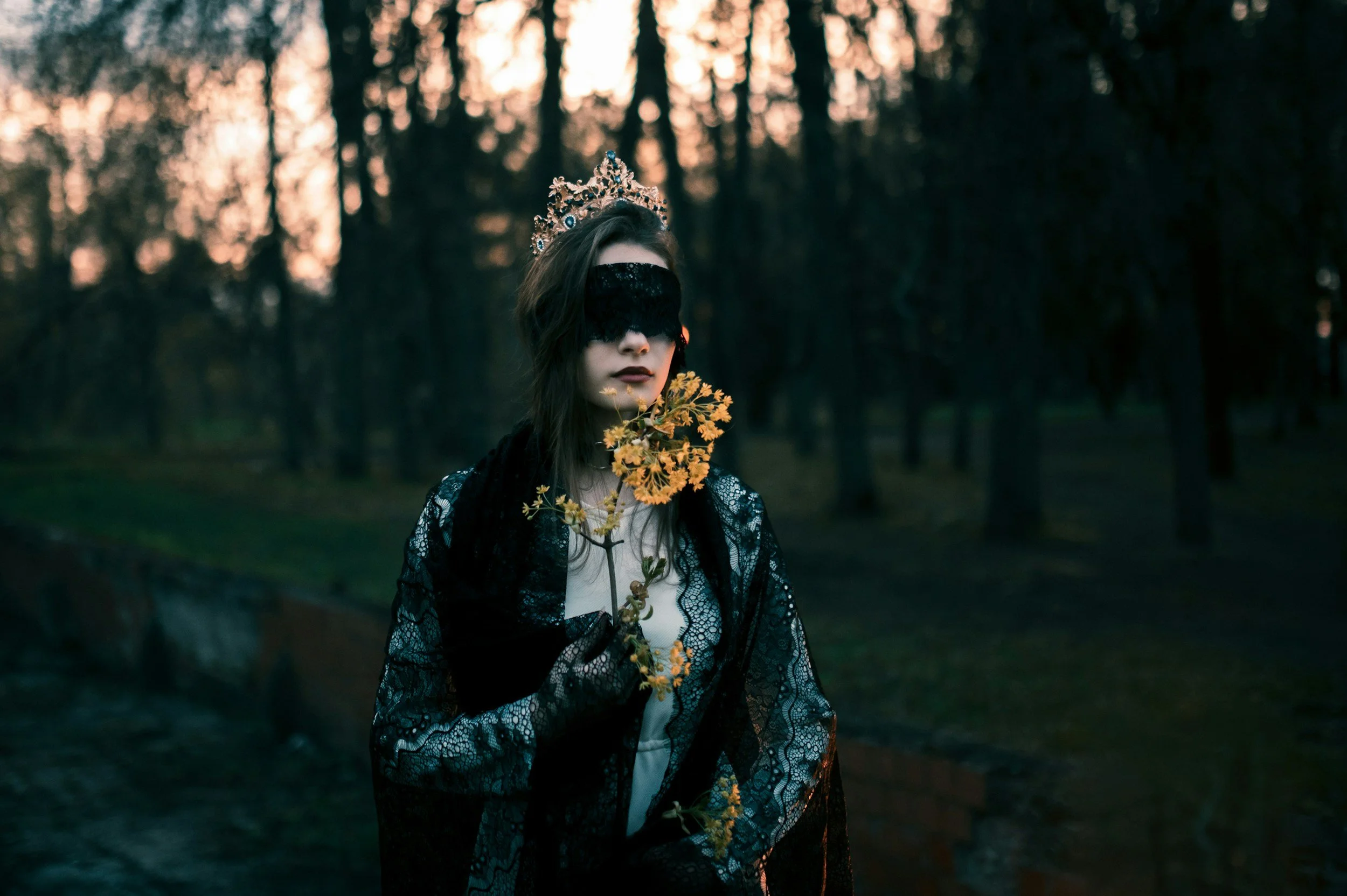 A woman wearing a black lace eye mask and a lace jacket stands outdoors at dusk, holding a yellow flower close to her lips with a blurred forest background.