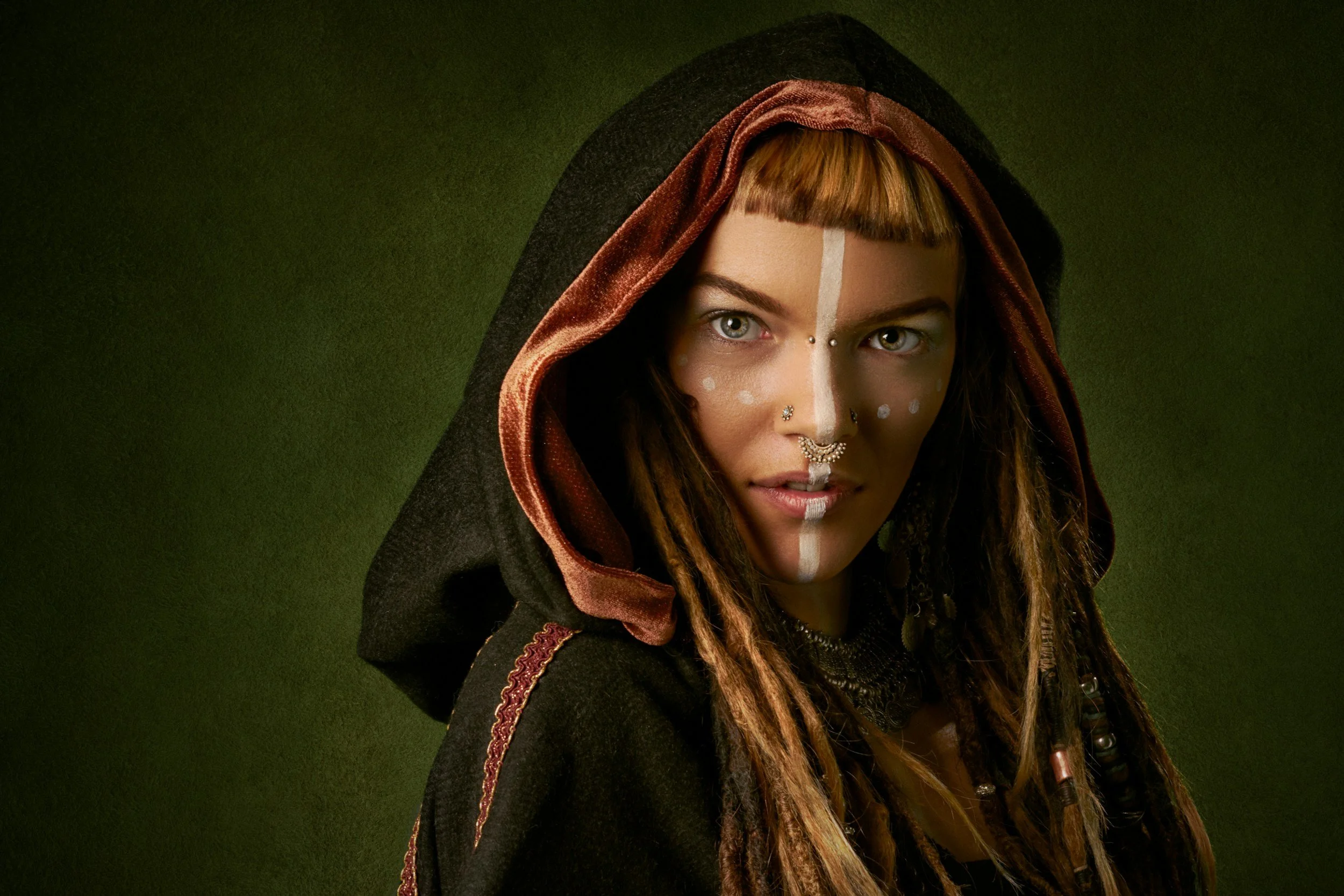 A woman with light skin and long dreadlocked hair, wearing a hooded cloak with a red and black lining, has face paint, jewelry including piercings and a nose ring, and is set against a green background.