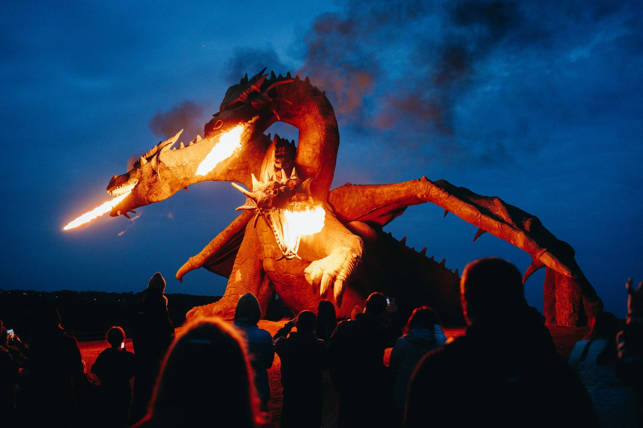 Large dragon sculpture with flames spewing from its mouth at dusk, crowd of people watching.