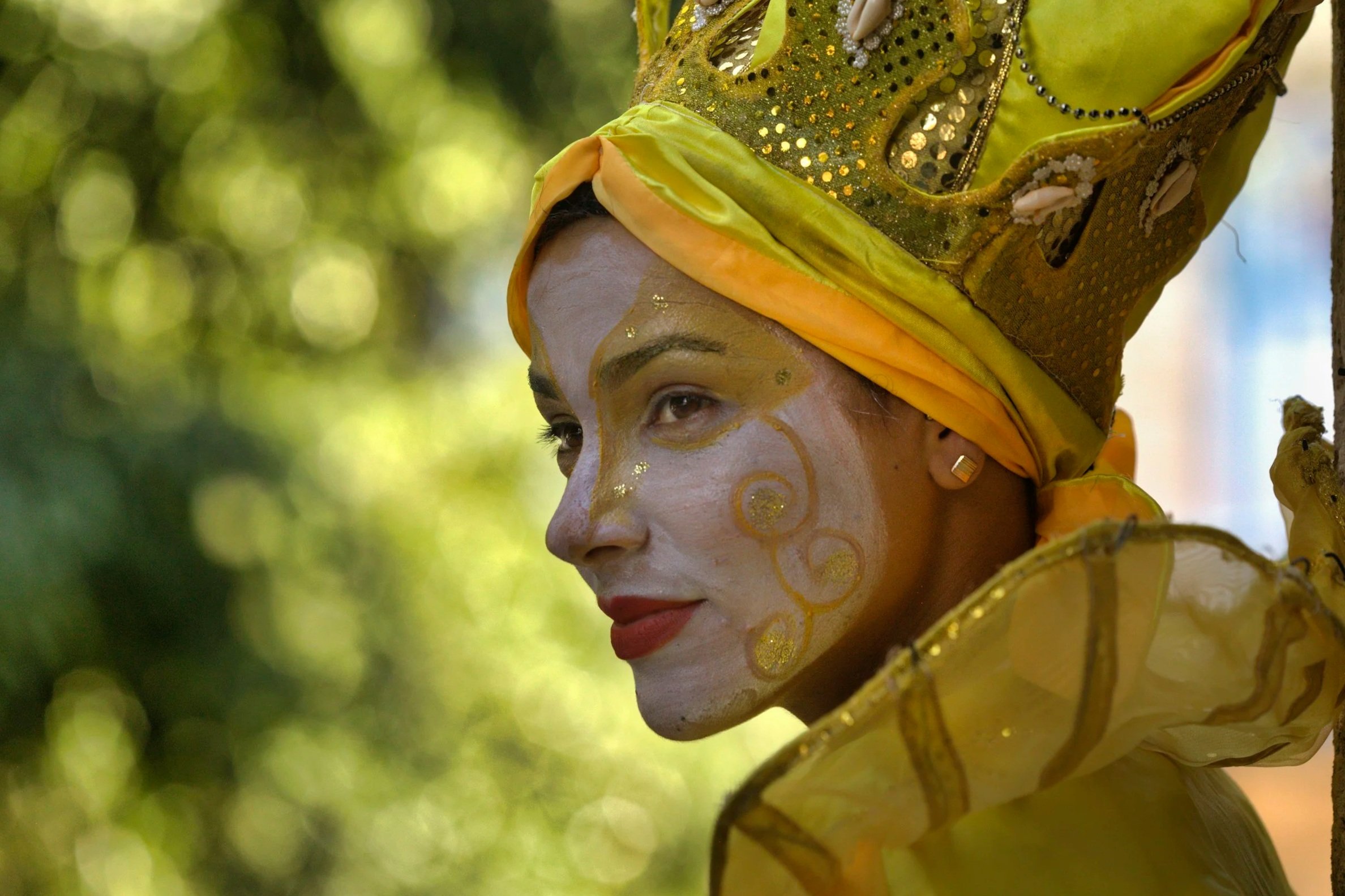 A person dressed in a vibrant yellow costume with gold and jewel embellishments, wearing a matching head wrap, and face paint with gold accents, during a cultural or traditional celebration outdoors.
