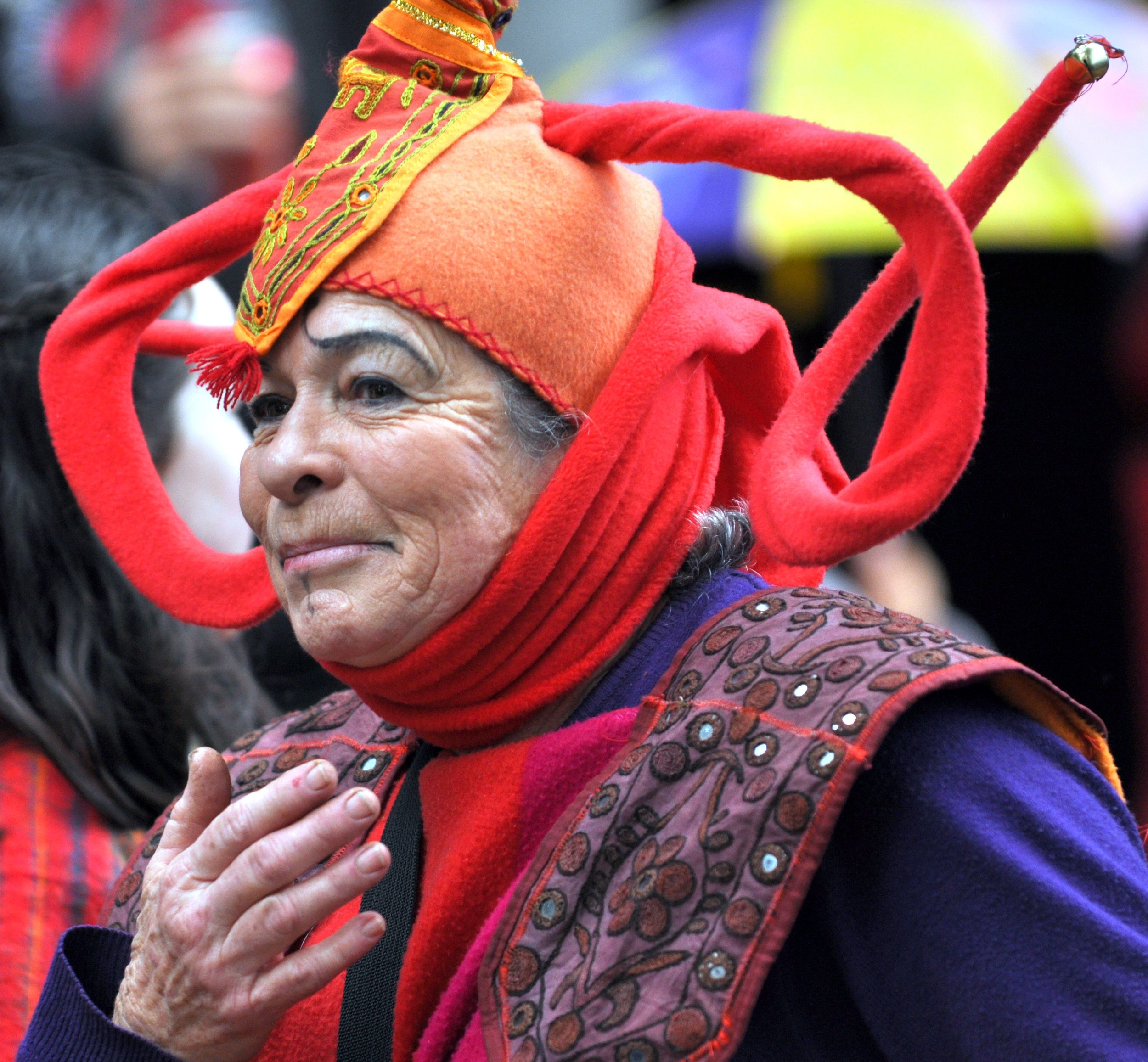 Close-up of a person wearing a colorful, ornate hat with embroidery, and a red clothing accessory with a pattern, hugging someone.