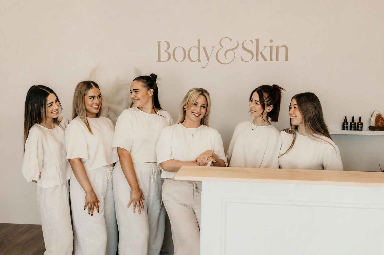 Group of six women smiling and talking at a reception desk in a wellness or beauty spa, with a sign that reads 'Body & Skin' on the wall behind them.