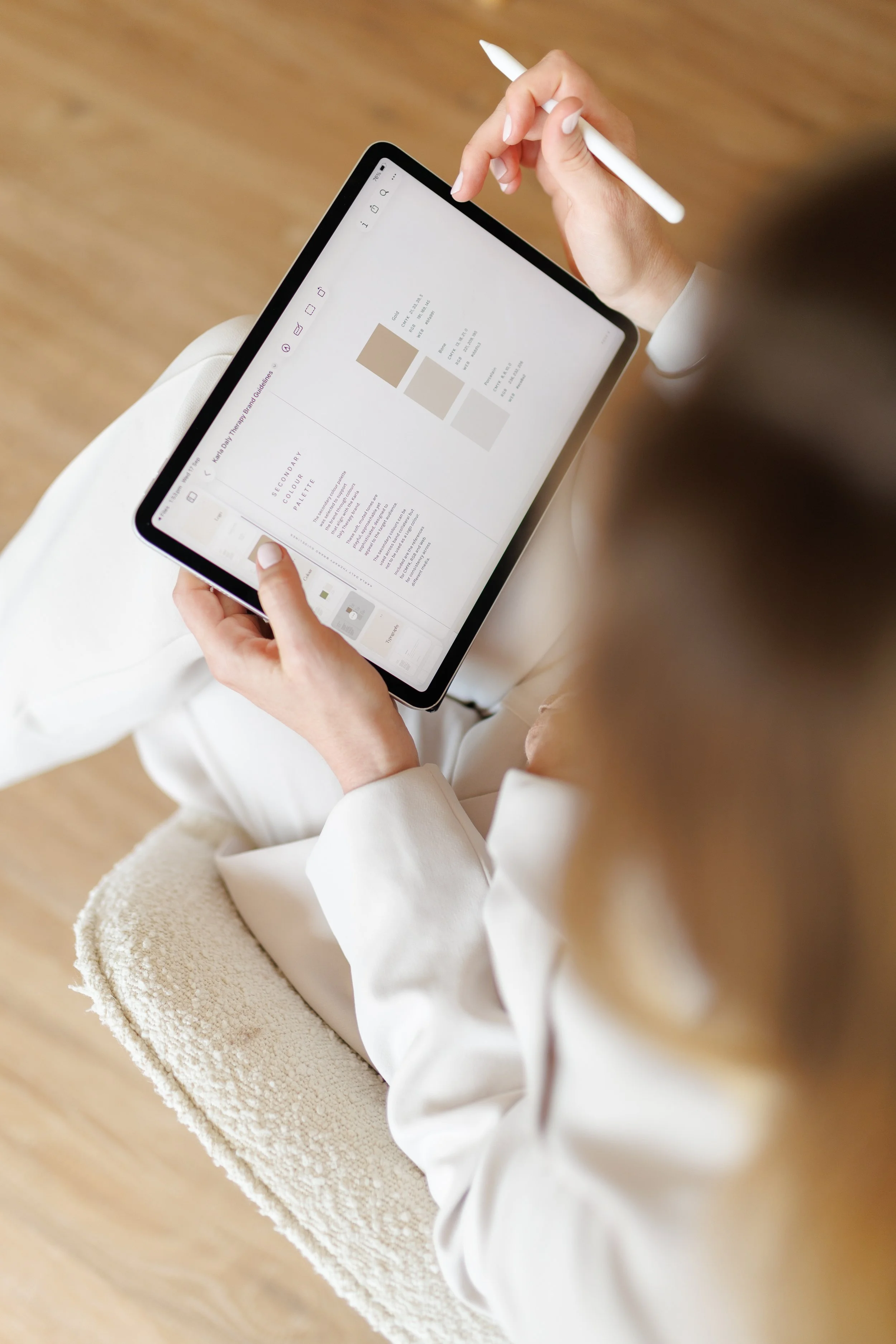 A person holding a tablet with a stylus, wearing a white suit, sitting on a textured beige chair with a wooden floor background.