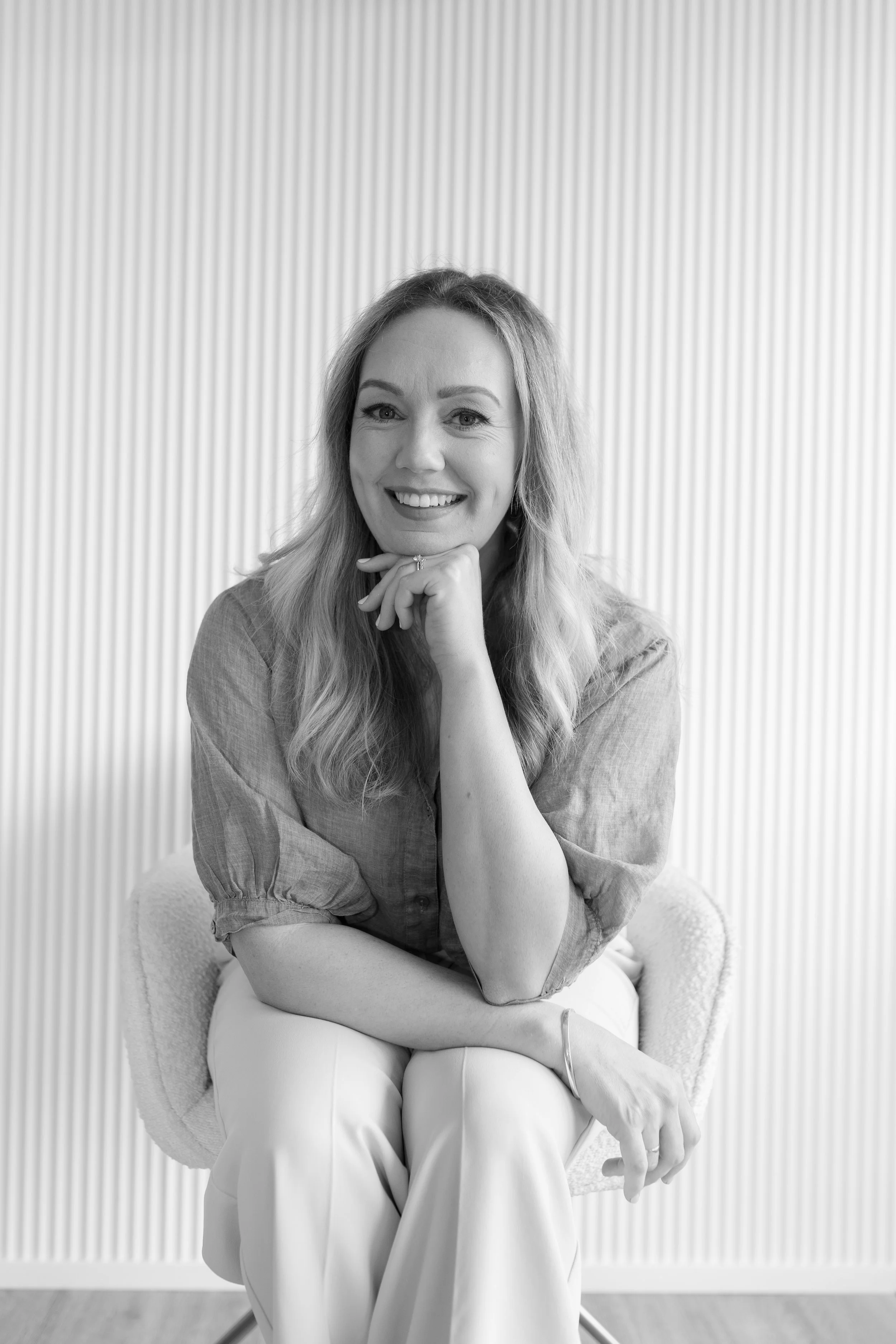 Black and white photo of a woman sitting on a chair with a relaxed pose, smiling, with one hand under her chin and the other resting on her lap. She has wavy hair and is wearing a long-sleeved top and white pants, with a plain, vertically striped wall in the background.