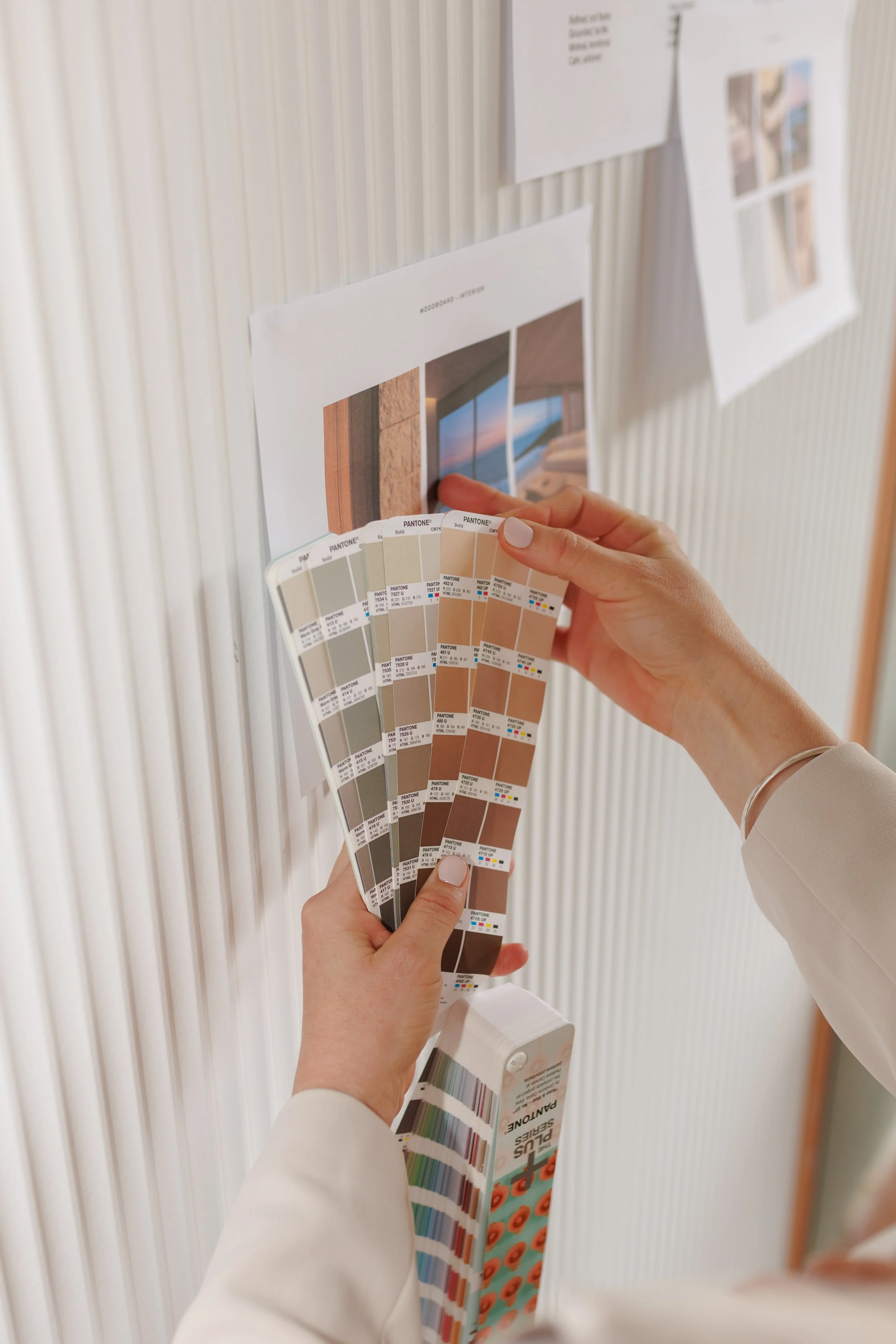 A person holding color swatch samples and reviewing interior design photos on a wall with a white textured background.