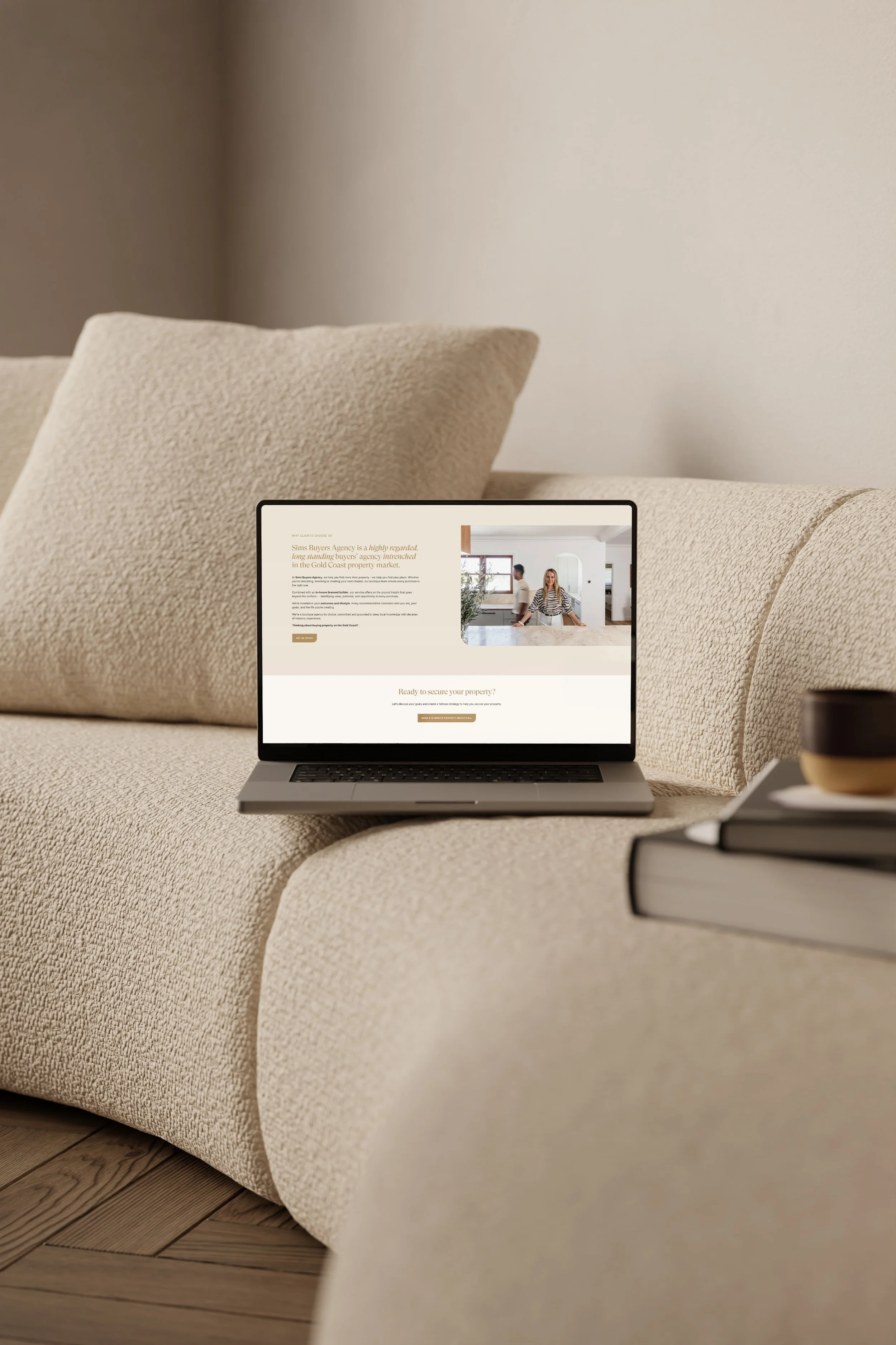 A beige textured couch with a beige pillow, a laptop on the armrest displaying a website with people, and a stack of books with a cup on top in the foreground. The background shows a plain wall and part of a wooden floor.