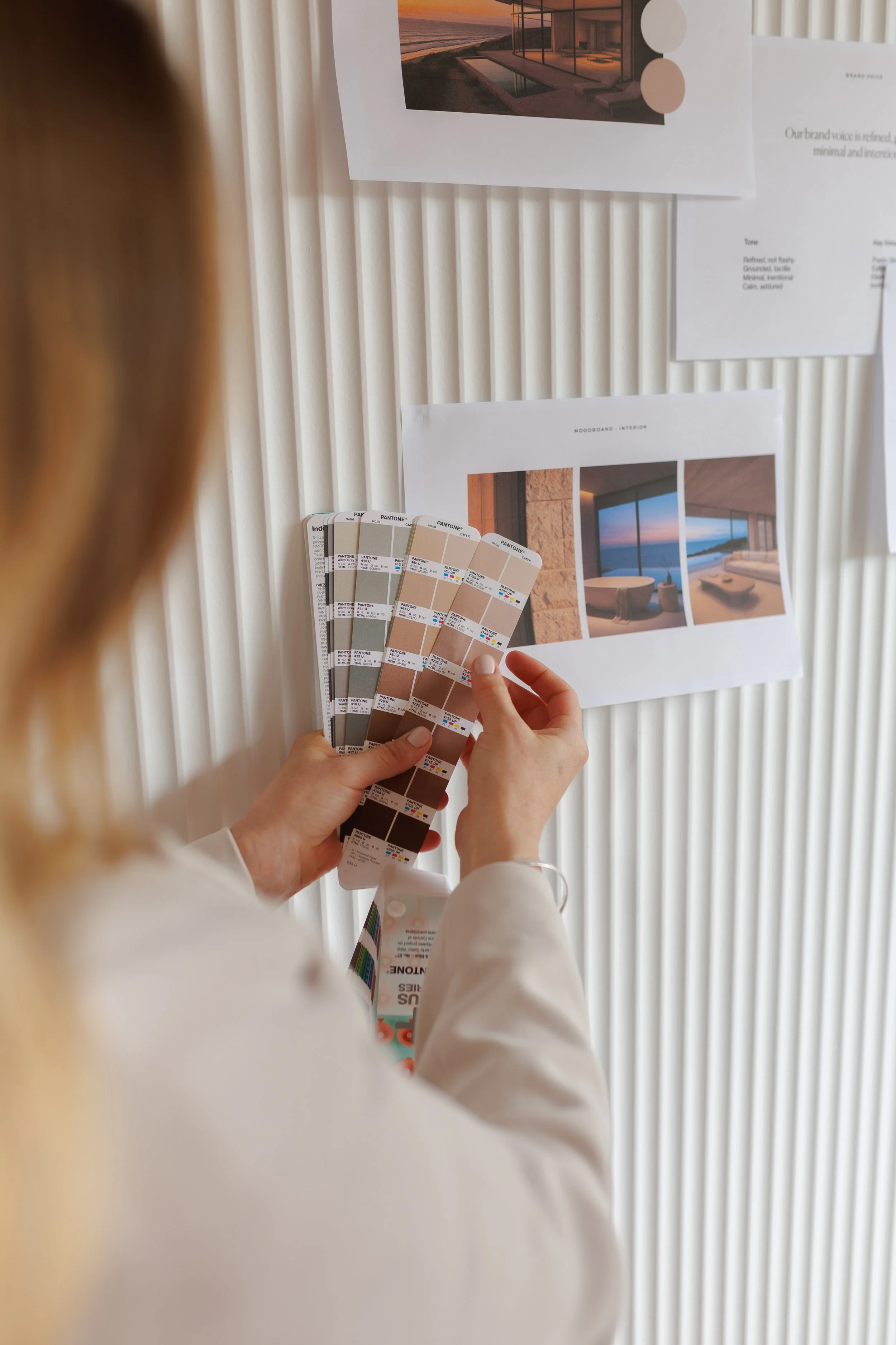 A person holding a palette of Pantone color swatches while examining interior design mood boards pinned on a wall, which feature images of modern interiors with ocean views and a sunset.