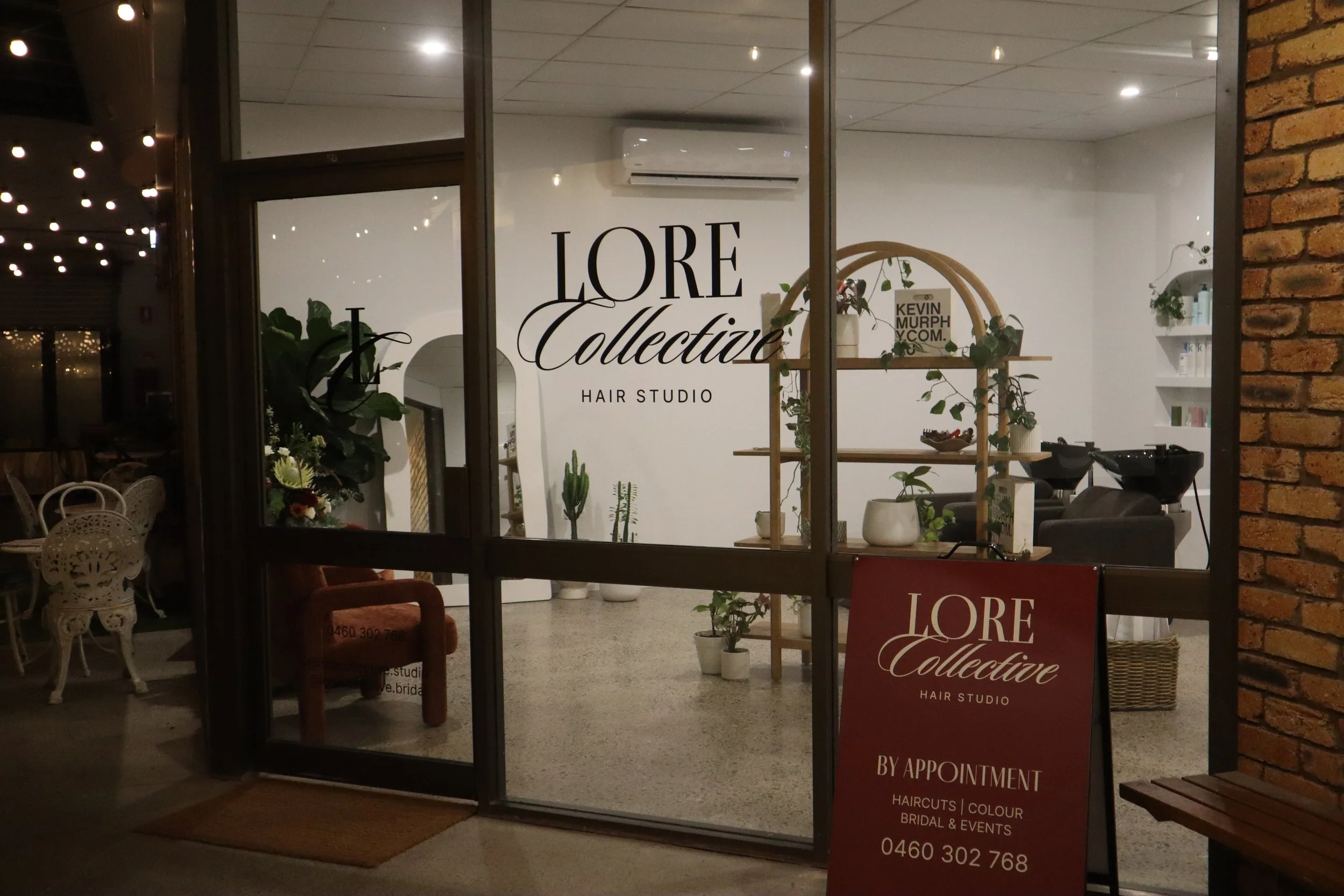 View through glass door showing a modern hair studio with white walls, wooden shelf with plants, and salon chairs inside. There is a sign for 'L'Oreal Collective Hair Studio'.