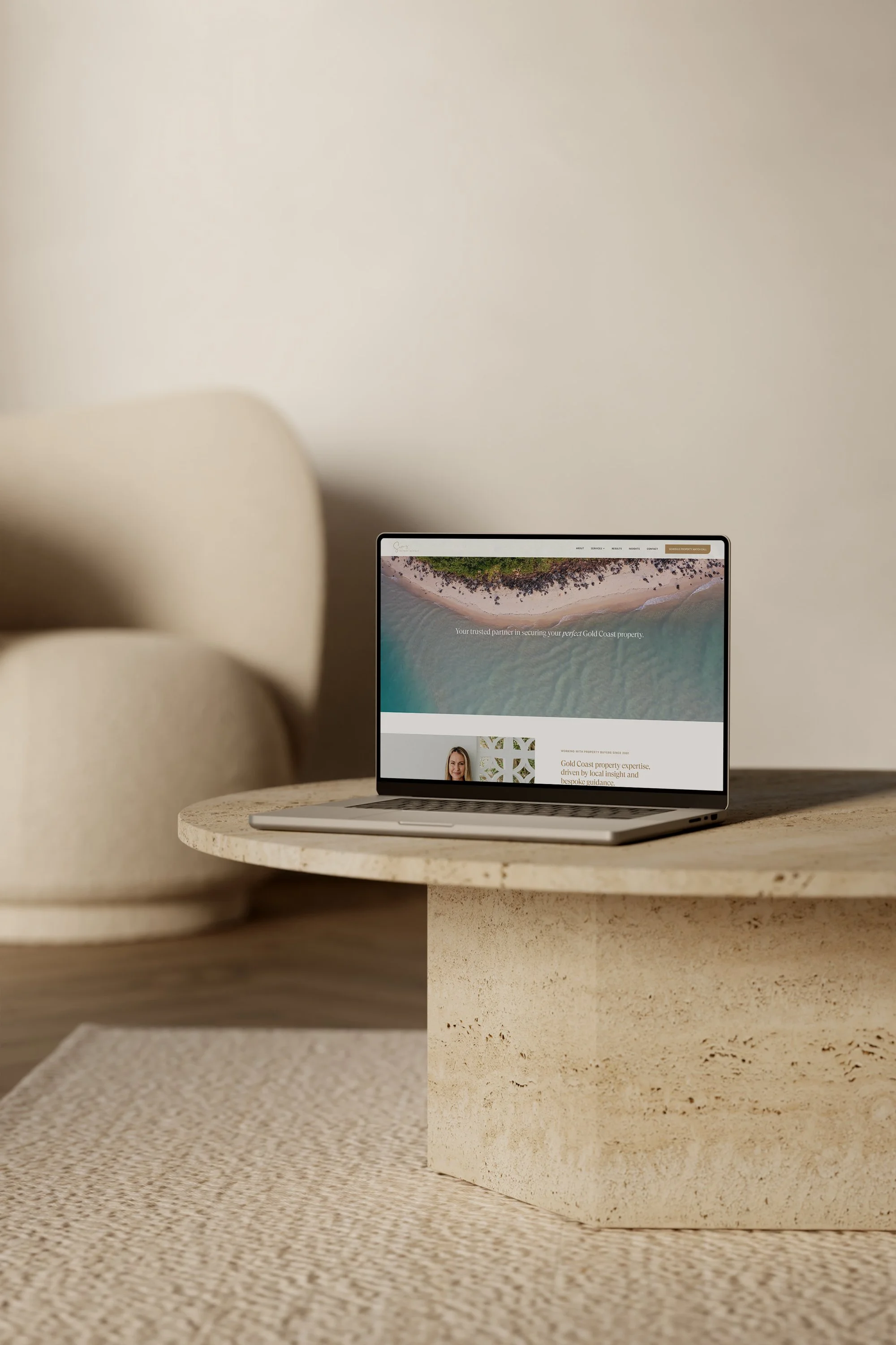 A laptop displaying a webpage with an aerial view of a beach with waves and green land, placed on a textured table near a beige textured divider or furniture.
