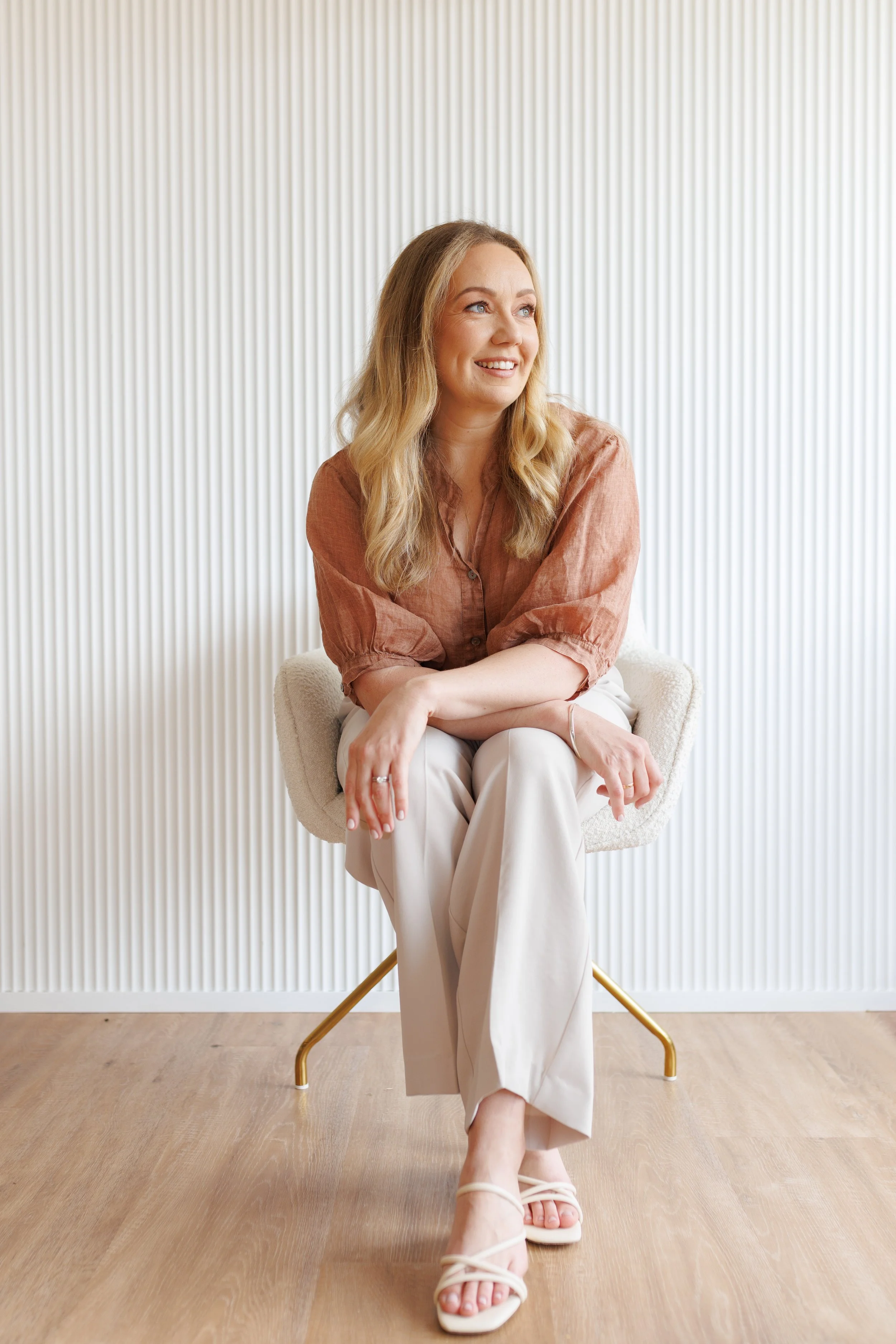 A woman with long blonde hair, wearing a brown blouse and cream-colored pants, sitting on a chair with a white background and wooden floor.