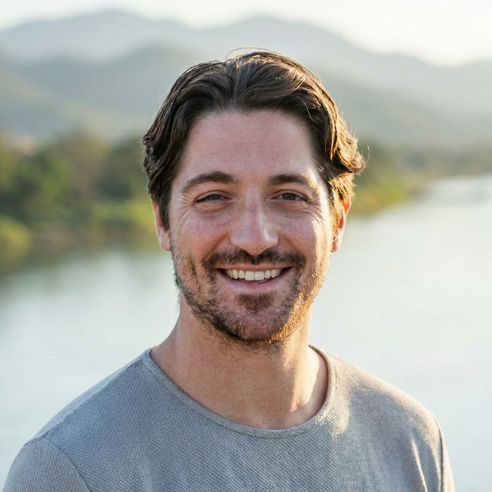 Headshot of Noah Zuckermann and wearing a light gray shirt, outdoors near water with mountains in the background.