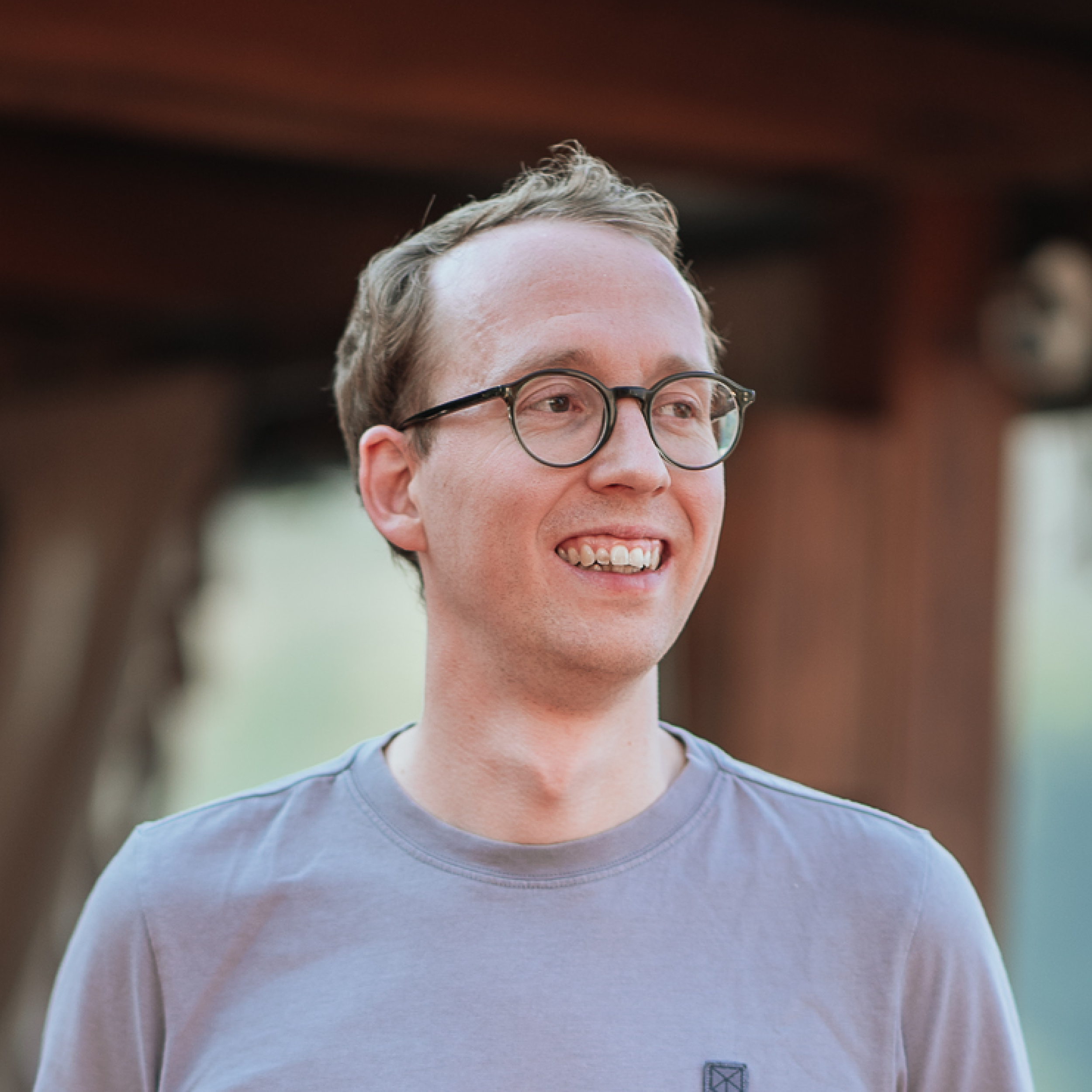 Close-up of Sascha Beele, wearing round glasses and a light gray t-shirt, smiling outdoors.
