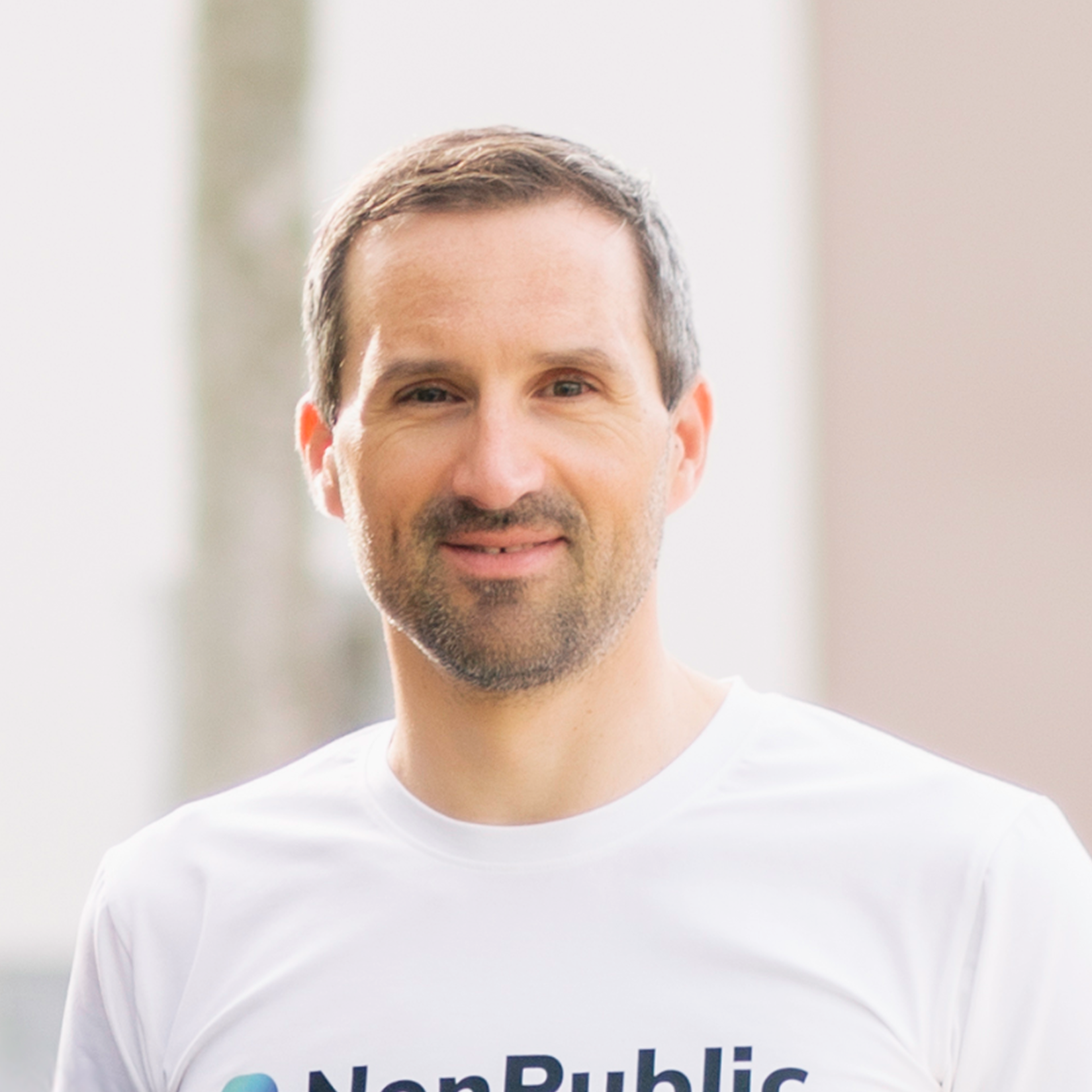 Close-up of Benni Lux, wearing a white T-shirt, standing indoors with a blurred background.