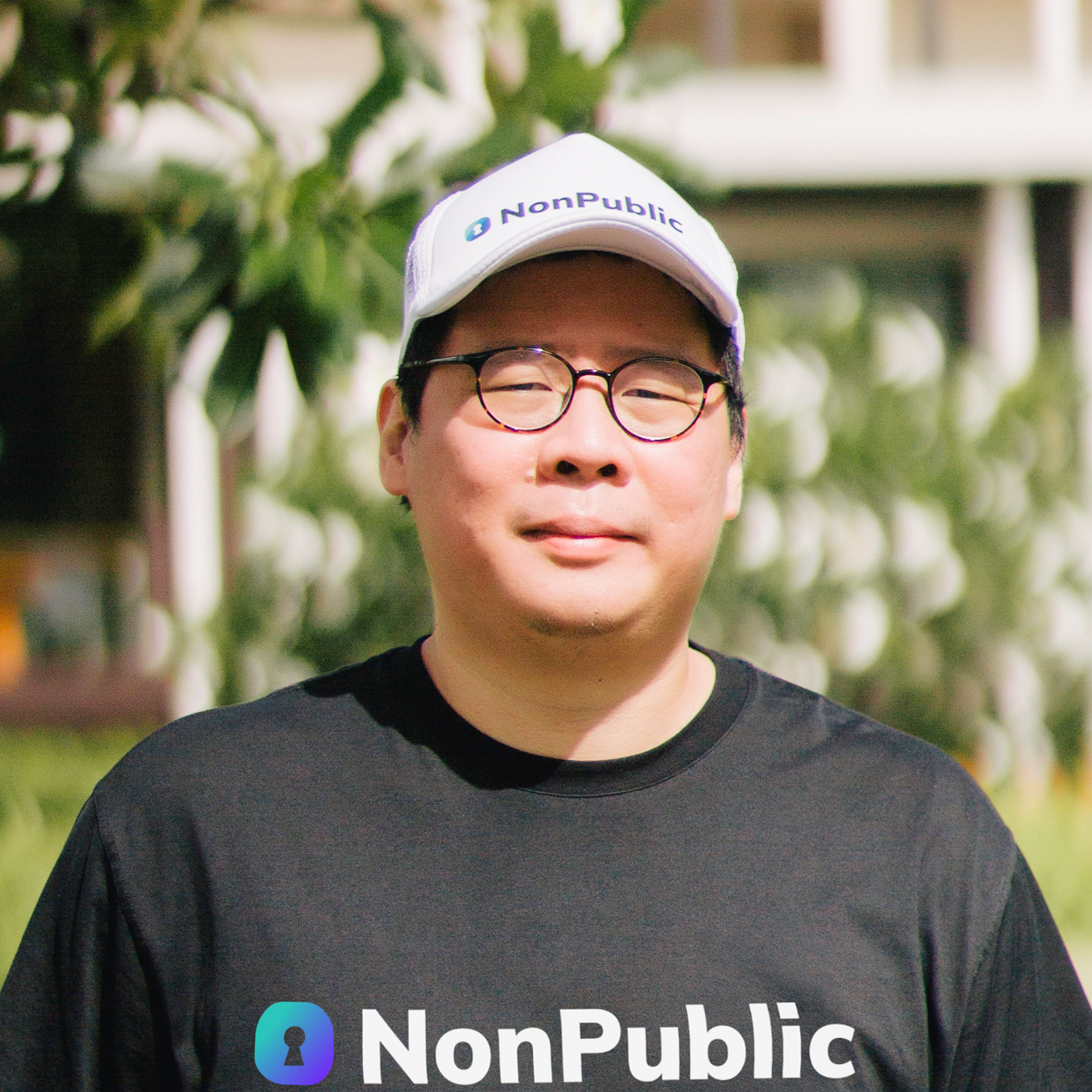 Close-up of a Ng Cheng Wei wearing glasses and a white cap with the logo and text of NonPublic, outdoors with blurred green foliage in the background.