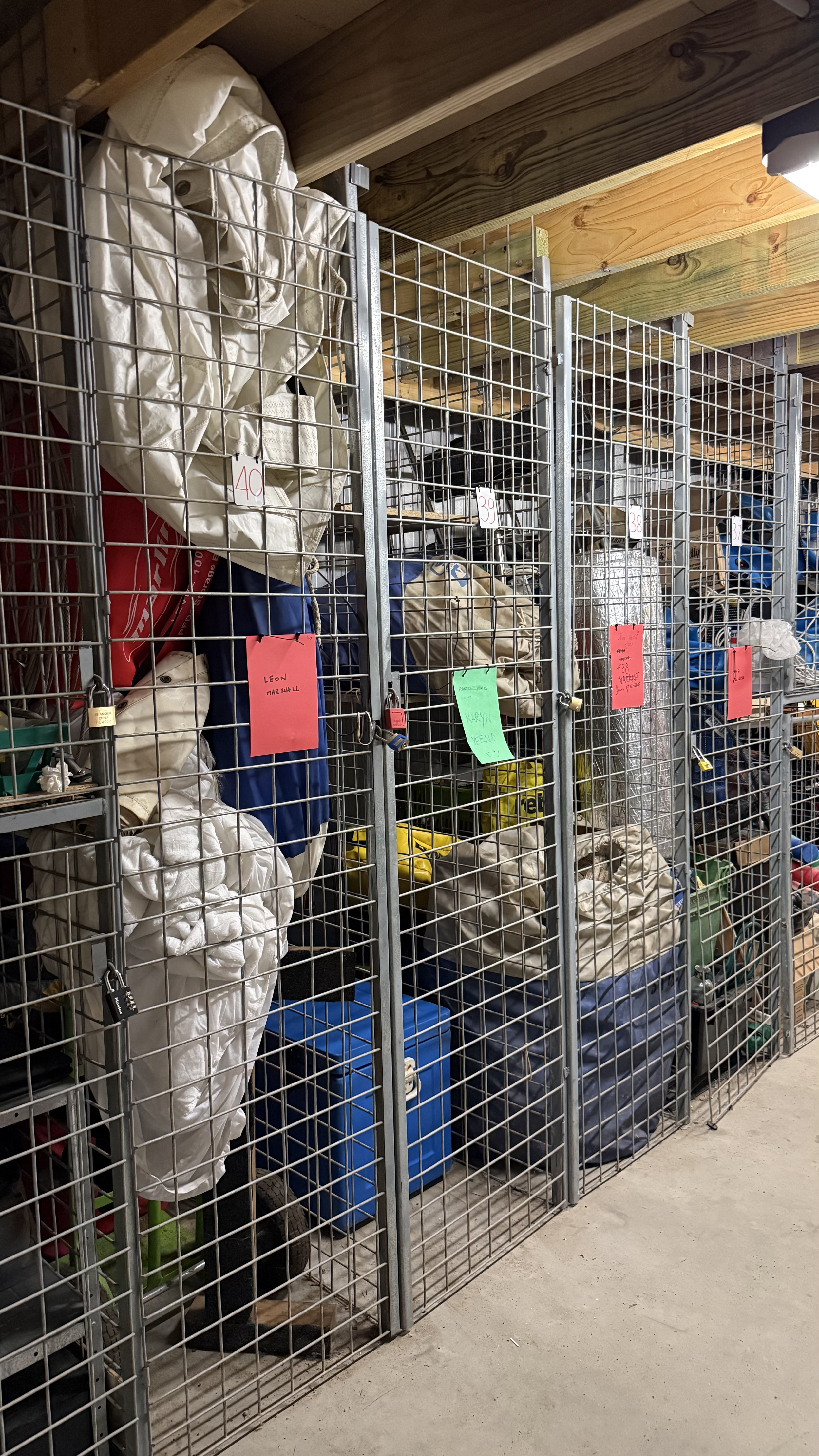 Storage lockers filled with closed, bagged, and bundled items, and some labeled with handwritten tags, in a secured metal cage, inside a wooden ceiling room.