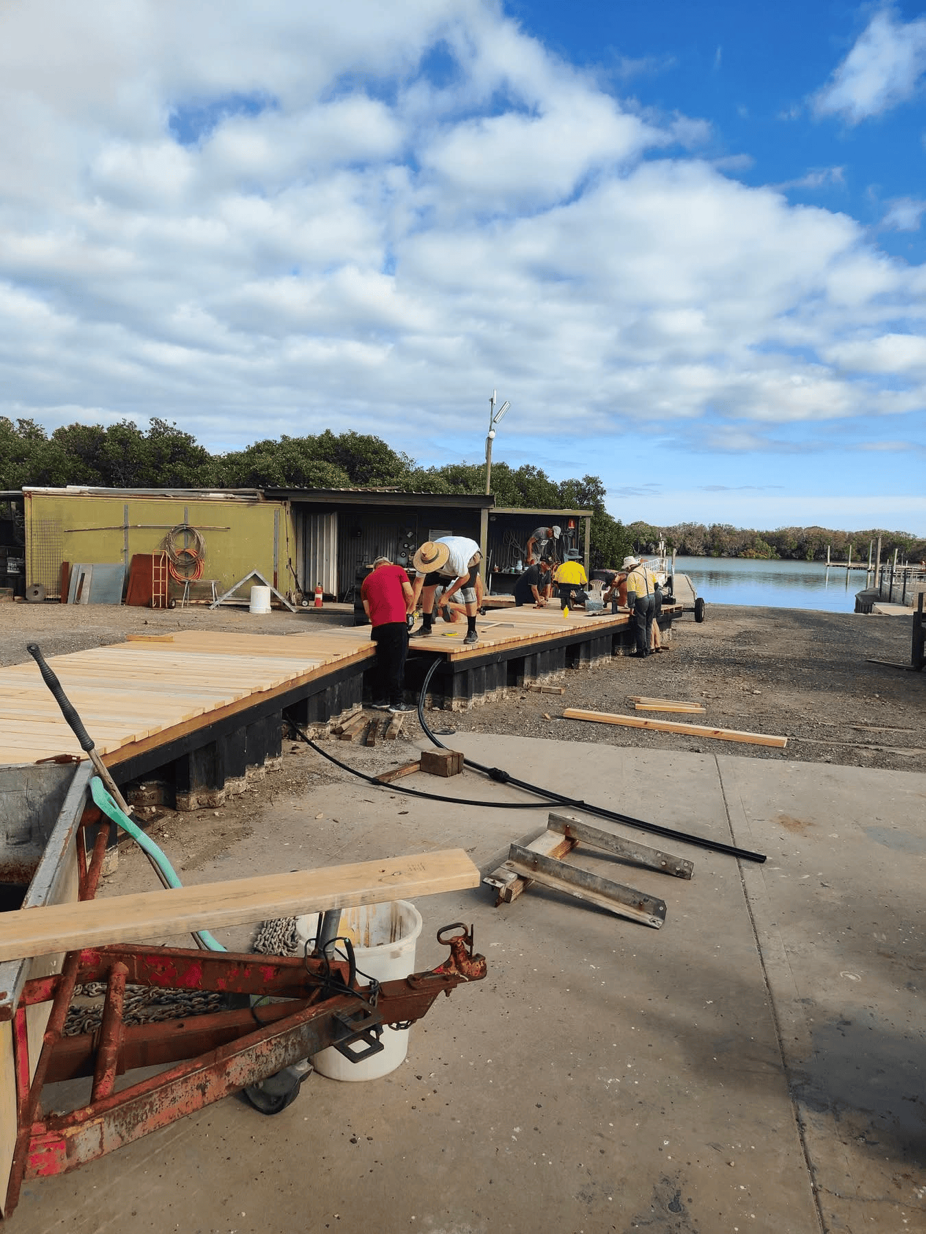 Workers building a wooden deck along a waterfront with a boat dock, with cloudy sky above and trees in the background.
