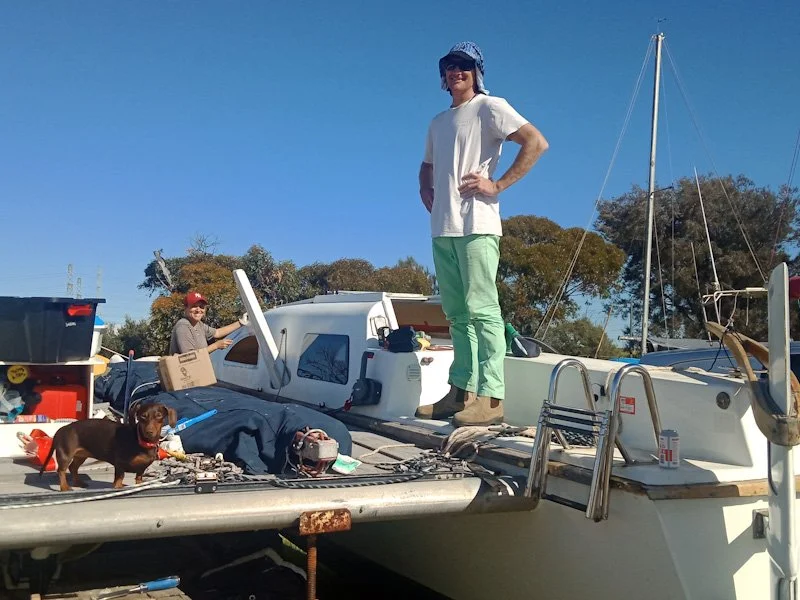 Two people and a dog on a boat docked at a marina under a clear blue sky, with trees in the background.
