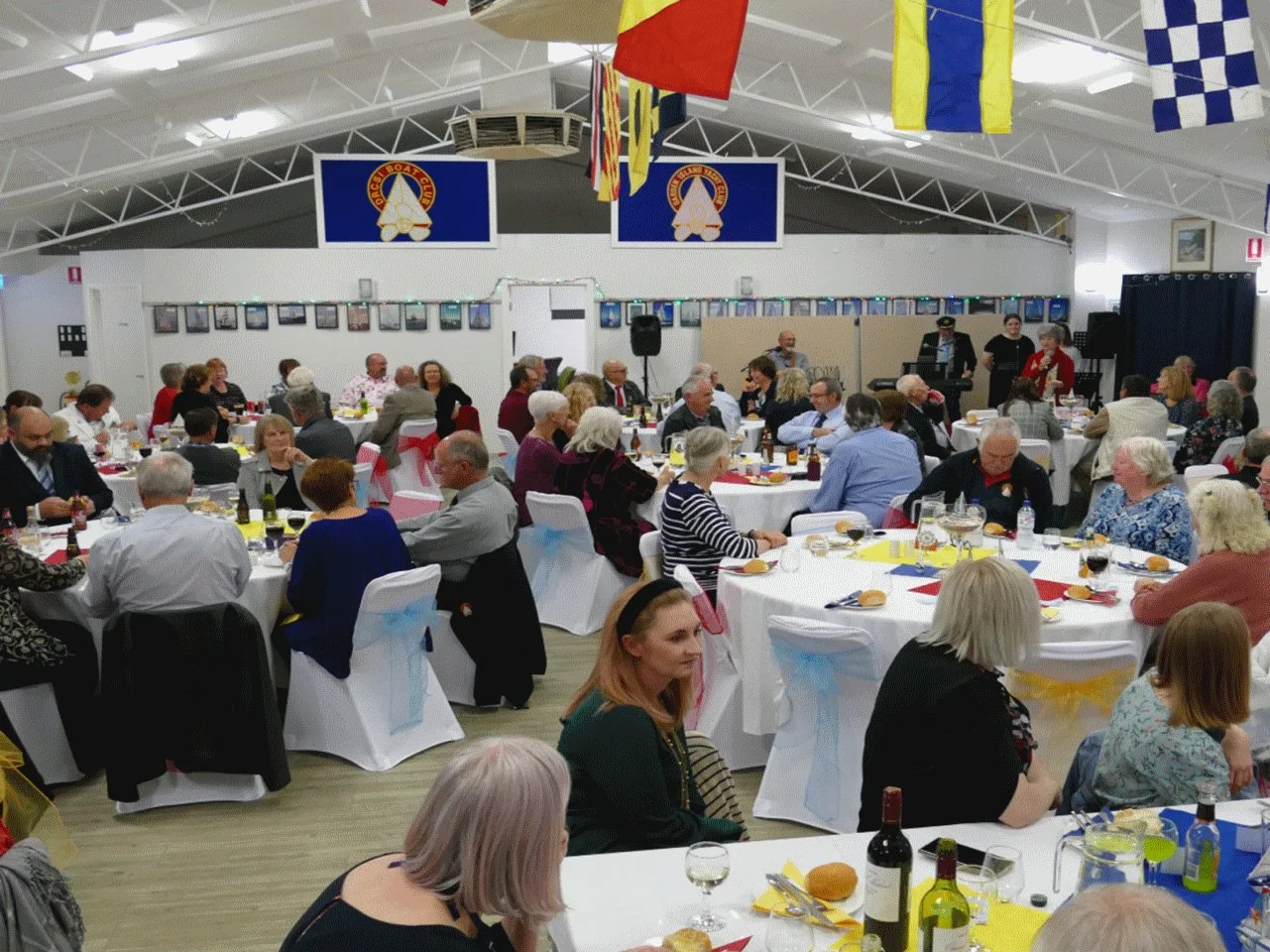 Large gathering of people seated at round tables during a social event or banquet in a decorated hall with flags hanging from the ceiling and framed photos on the wall. People are enjoying food and drinks.