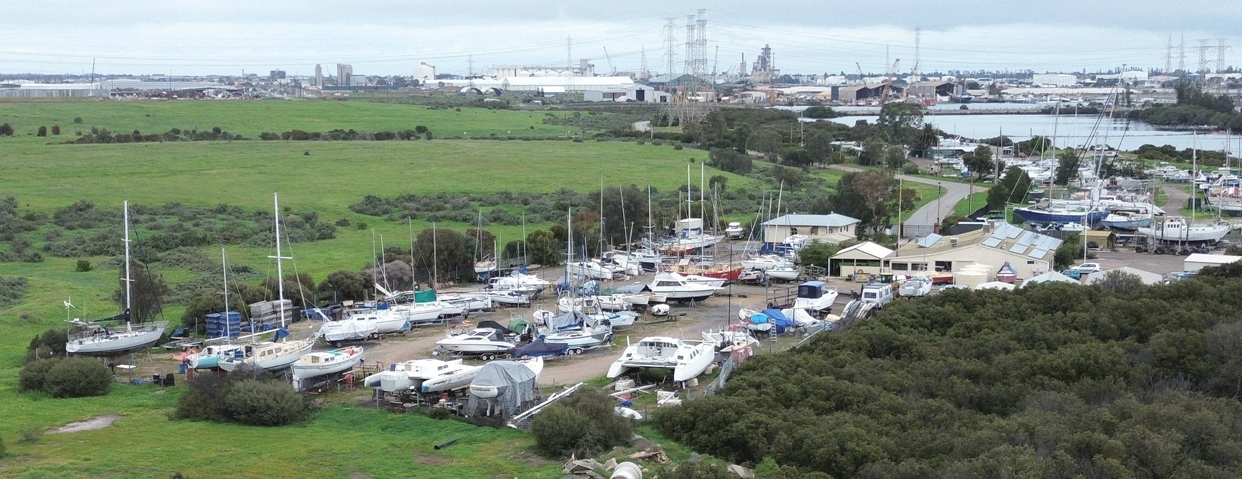 A boat storage yard with numerous sailboats and motorboats on land, surrounded by green fields and nearby water bodies in the background.