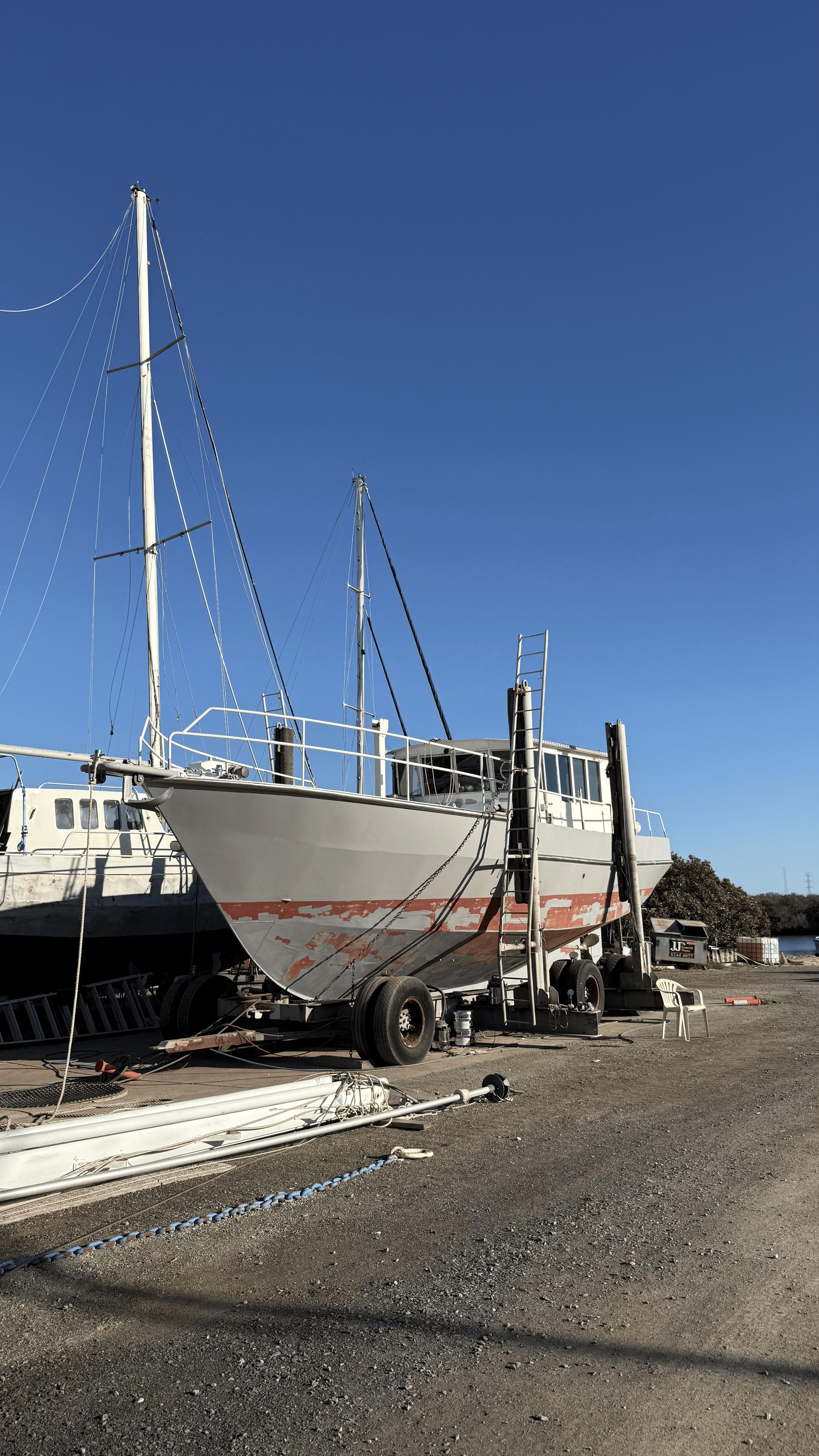 A boat on a trailer is parked on a gravel lot with other boats, equipment, and chairs around, against a clear blue sky.