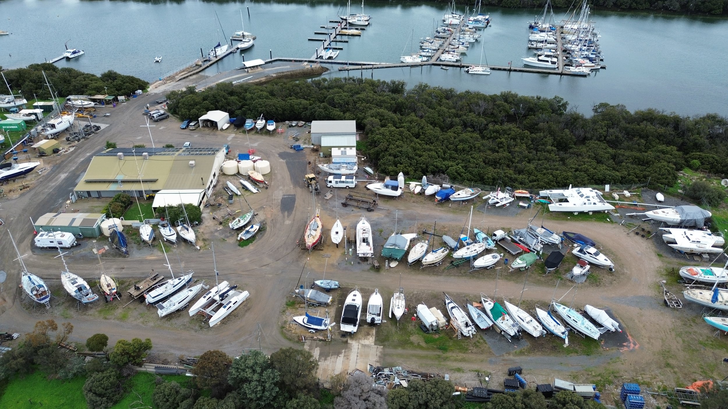 Aerial view of a marina with numerous boats docked in the water and on land, surrounded by trees and a building with equipment and vehicles nearby.