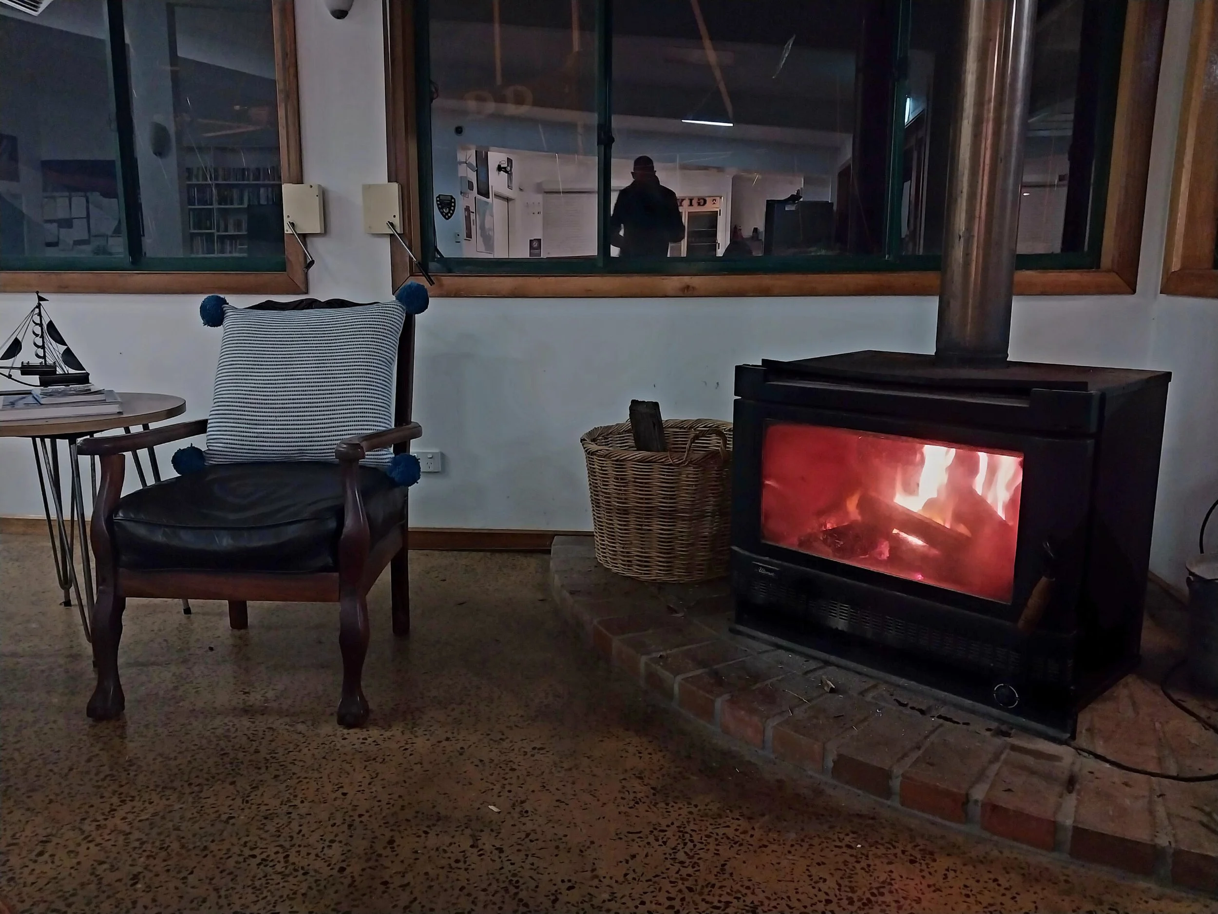 A cozy living room with a wood-burning stove, a wicker basket, a wooden armchair with a navy and white striped cushion, and a small side table with a decorative sailboat. Through the window, a person is visible inside a well-lit room, and a fire is burning inside the stove.