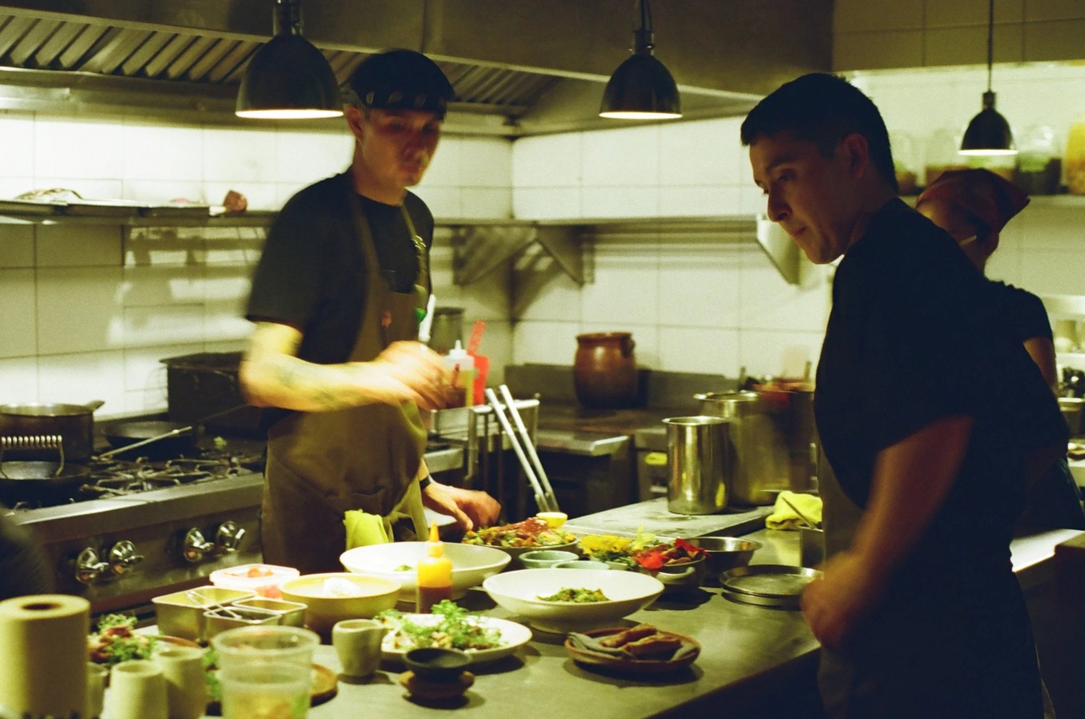 Two chefs in a kitchen preparing food with various bowls of ingredients on the counter in front of them.