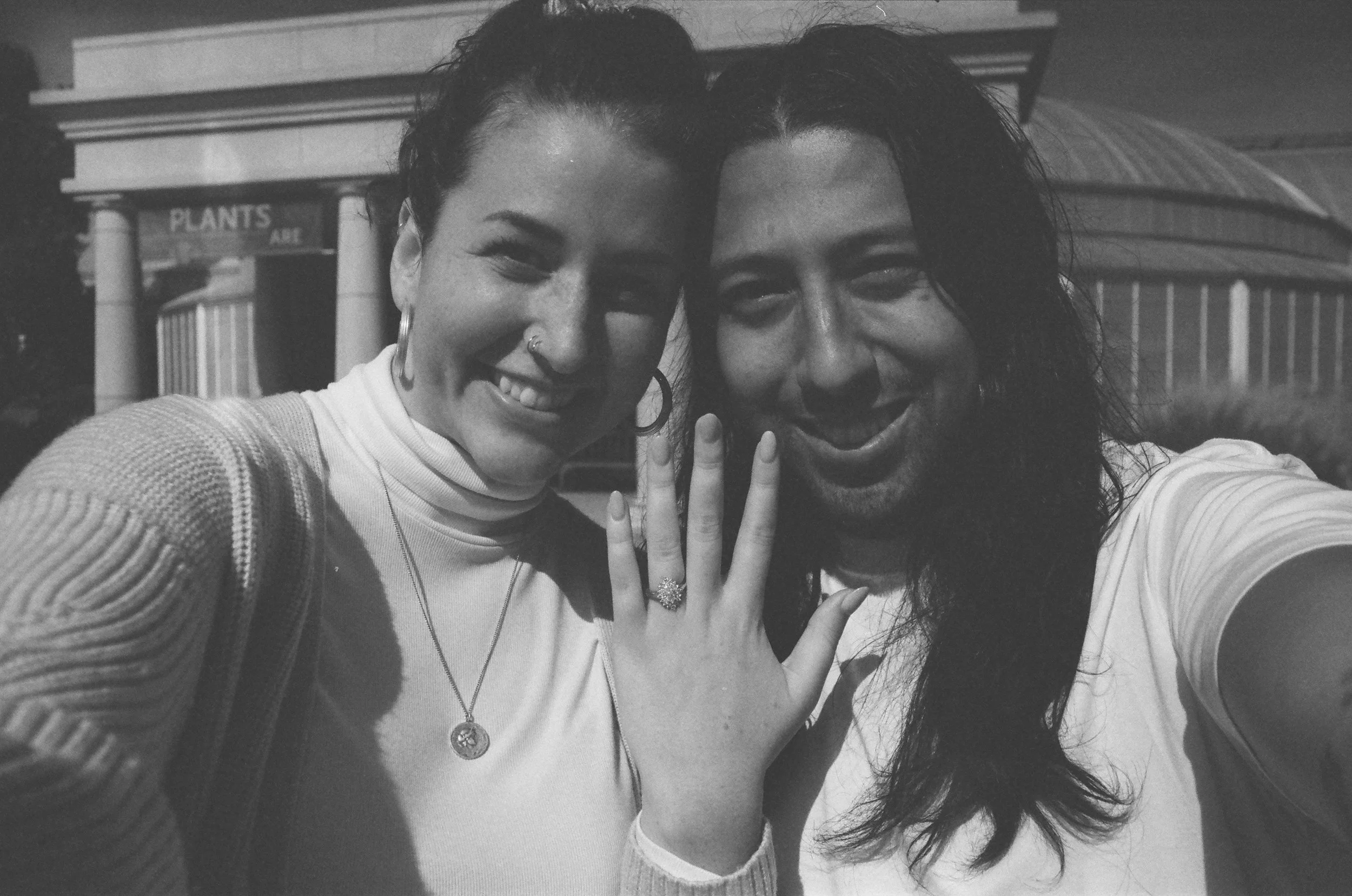 Two smiling women taking a selfie together, with one showing a peace sign, in front of a building with columns and a sign that reads 'PLANTS'.
