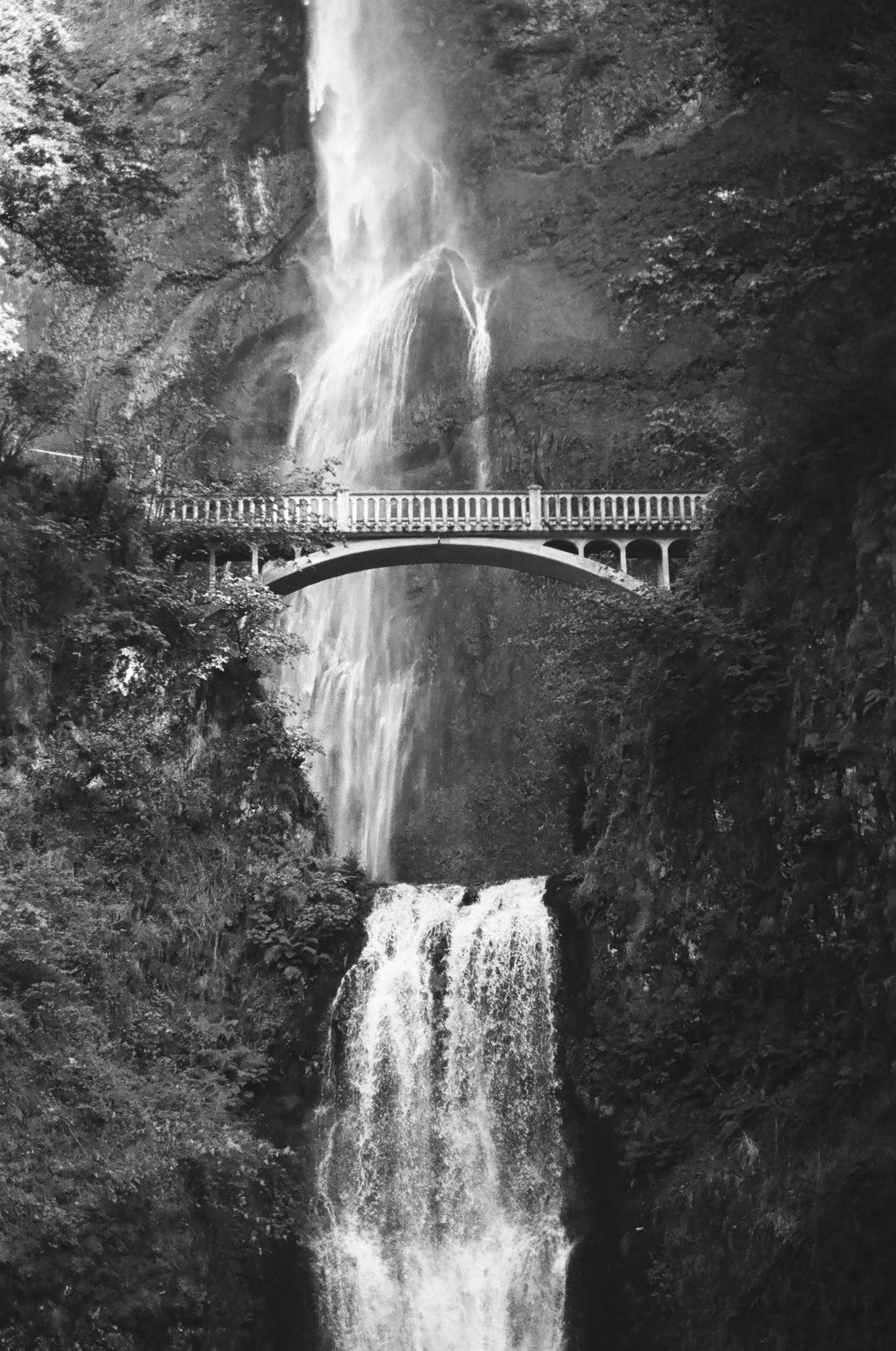 Black and white photo of a multi-tiered waterfall cascading down a rocky cliff surrounded by trees, with a bridge crossing over the water.