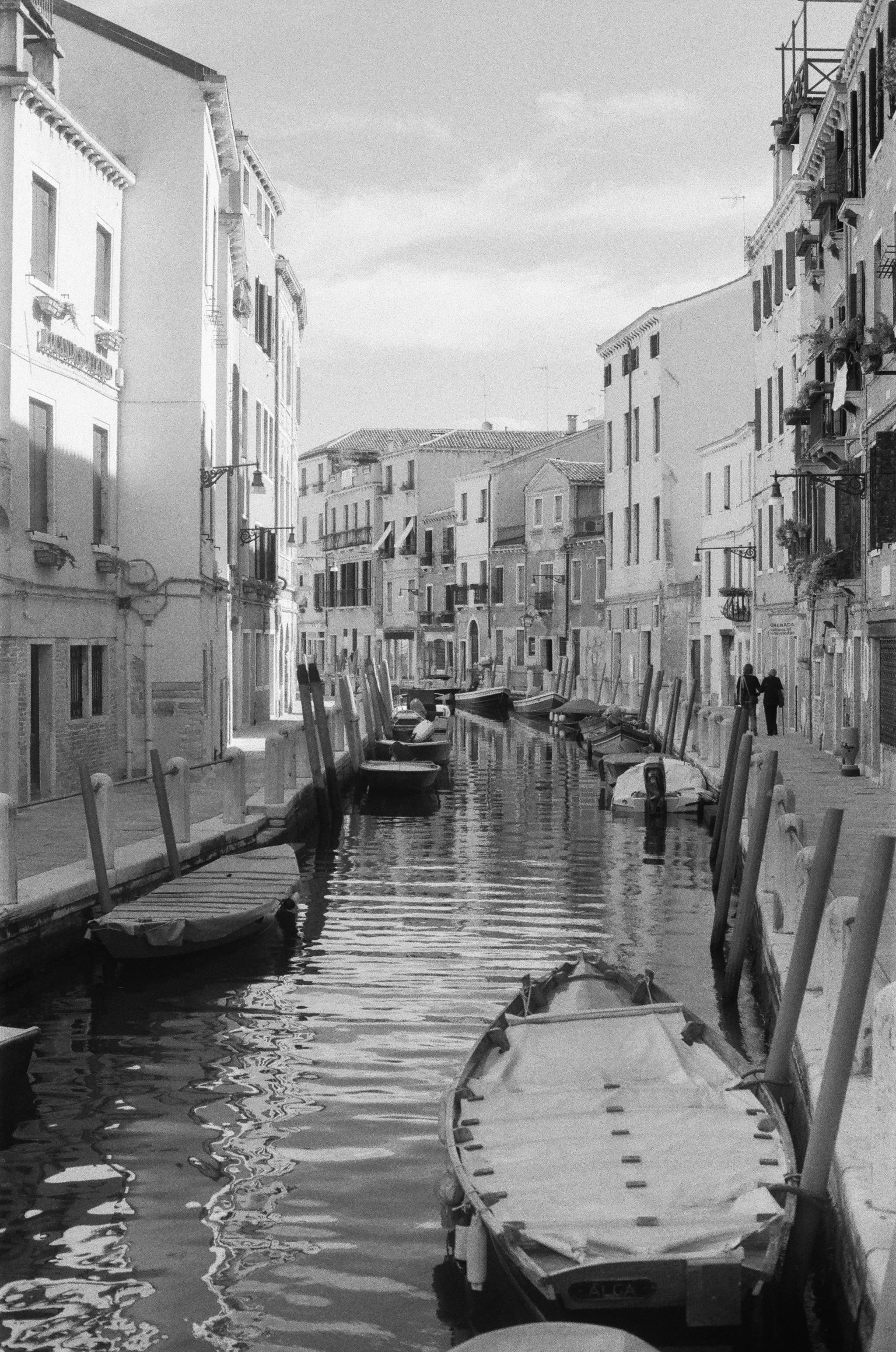 A canal lined with buildings on each side, boats docked along the edge, and a couple walking on the sidewalk.