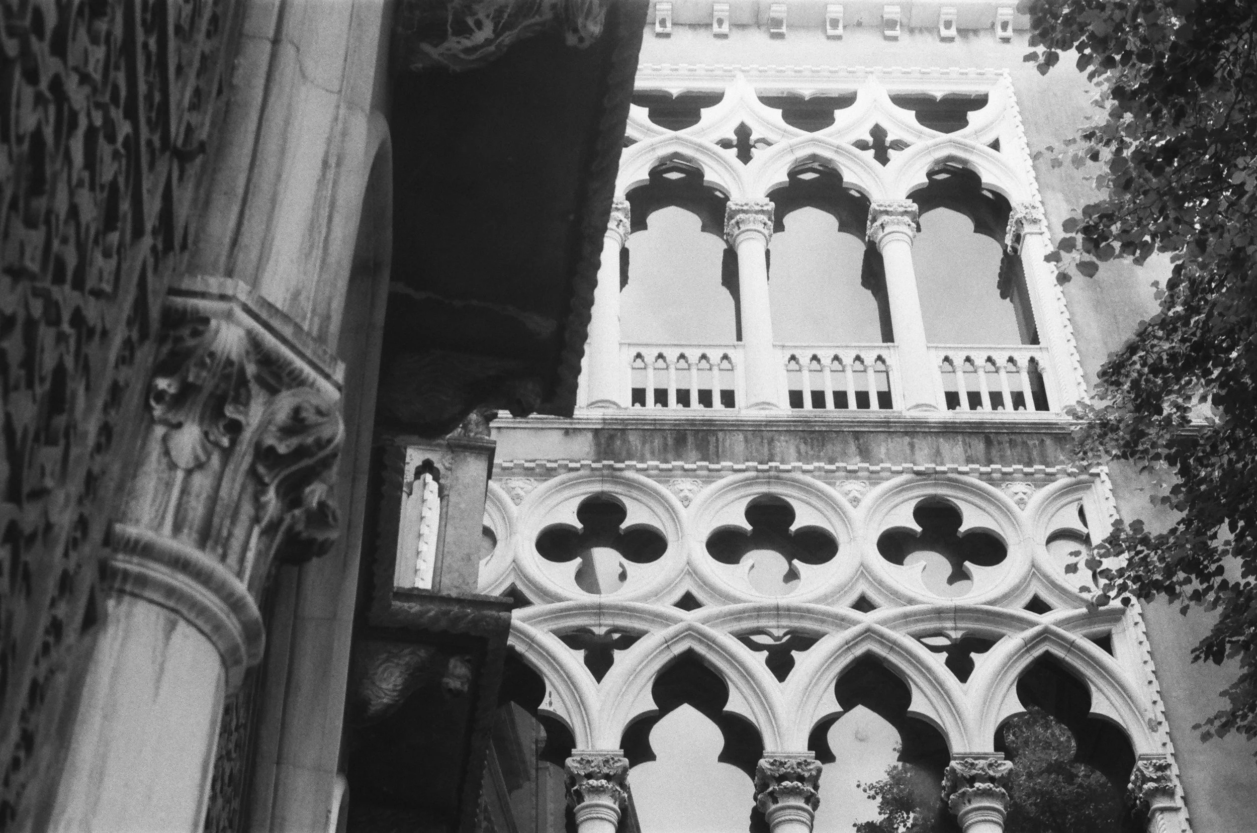 Black and white photograph of intricate Gothic-style architectural details, including pointed arches, columns, and decorative stone carvings, with a brighter building facade in the background and some tree foliage on the right.