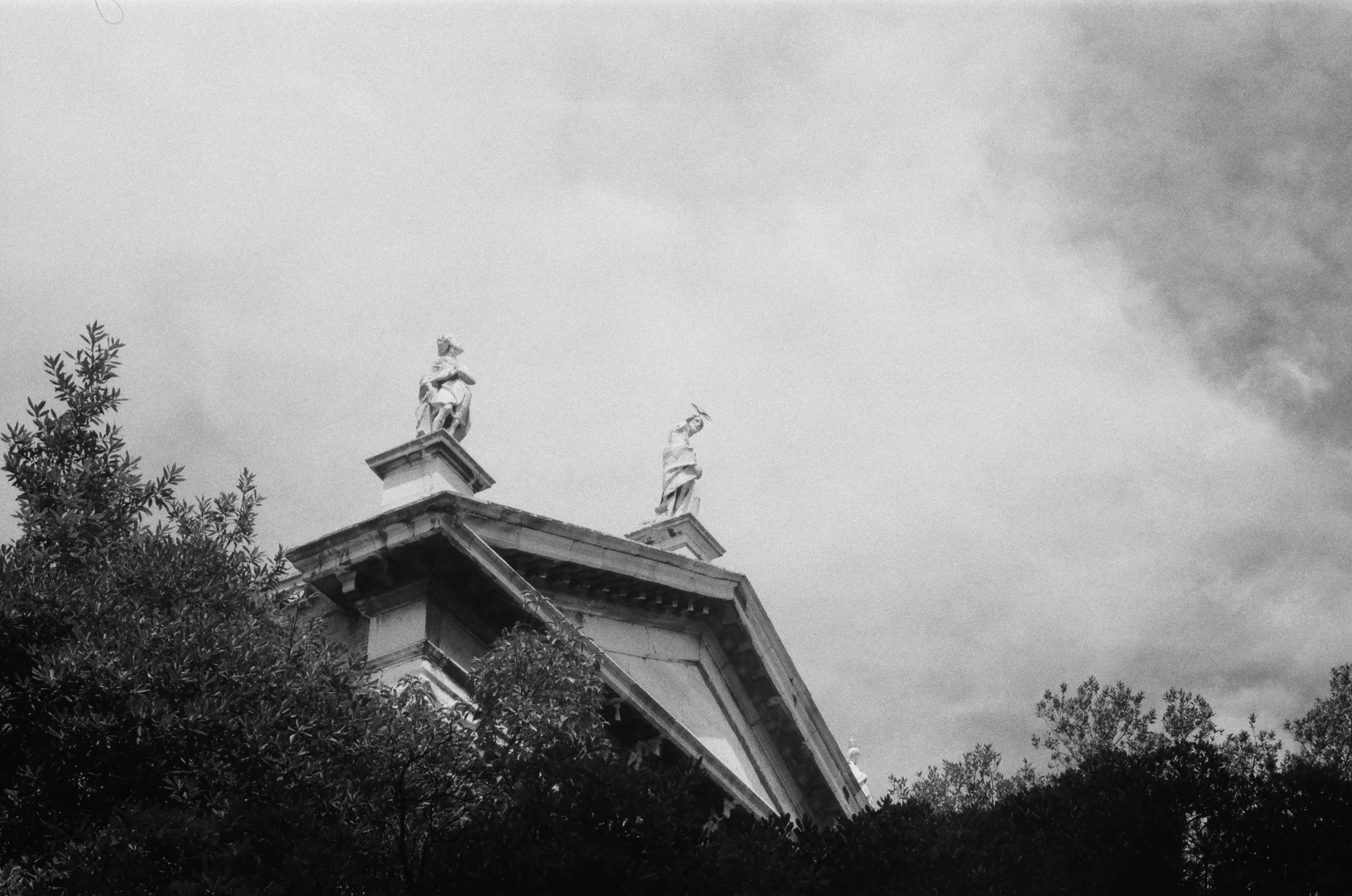 Black and white photo of a building with statues on top, surrounded by trees under a cloudy sky.