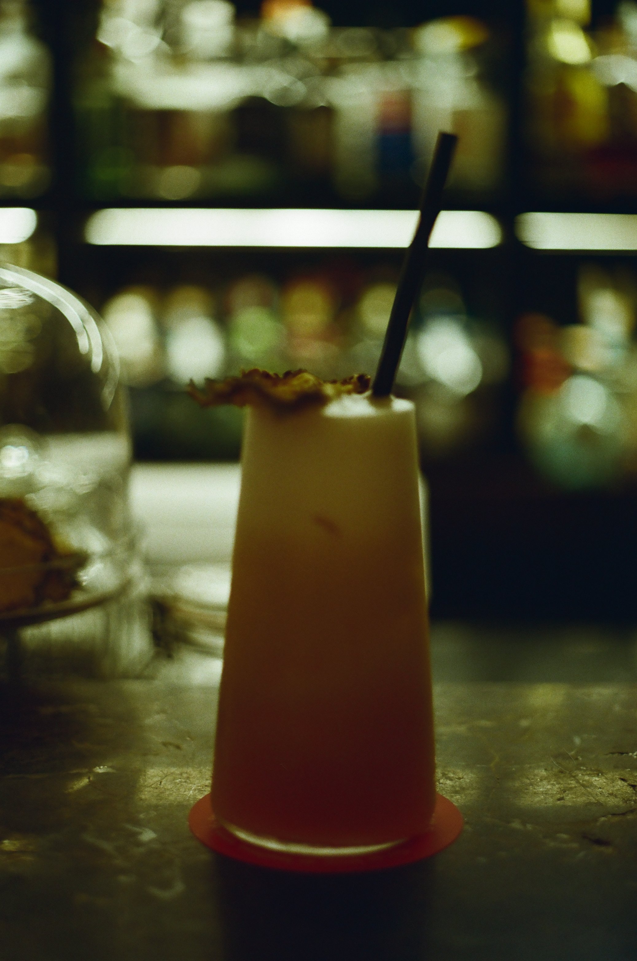 A small orange traffic cone with a black straw inserted on top, standing on a kitchen countertop. Blurred bottles and jars are visible in the background.