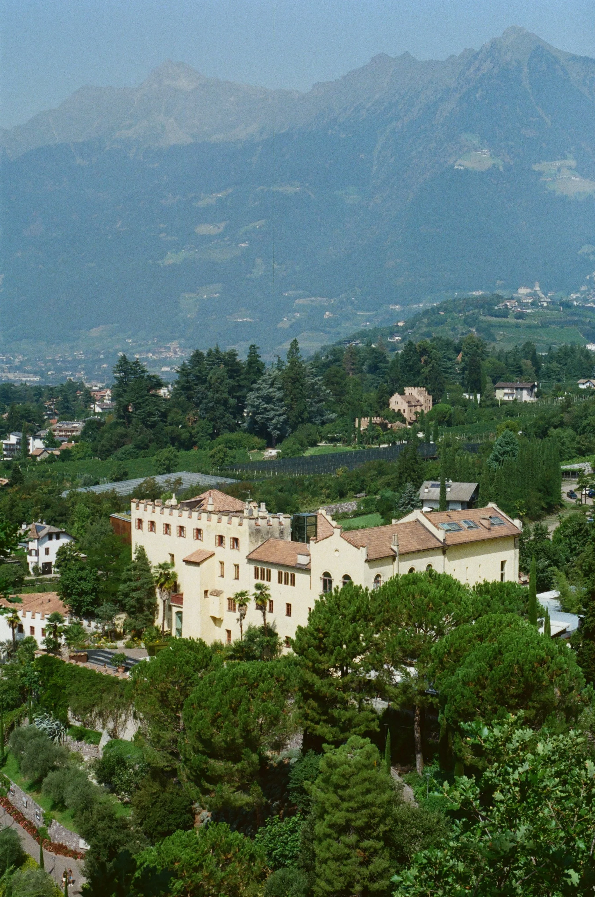 A scenic view of a hillside town with lush green trees, a castle-like building, and distant mountains in the background.