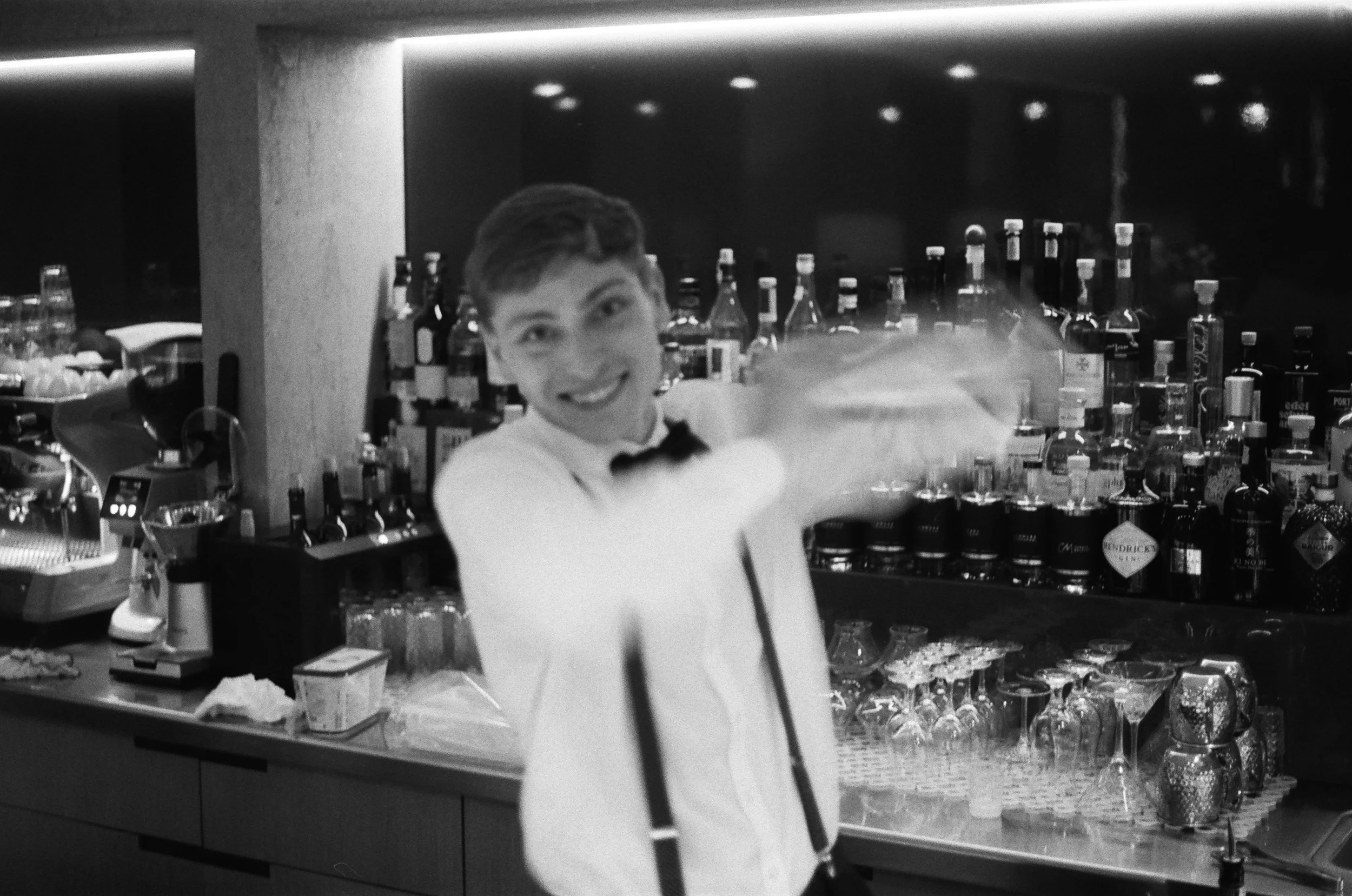 A smiling bartender in a white shirt and black bow tie serving drinks behind a bar with various bottles of alcohol on shelves.