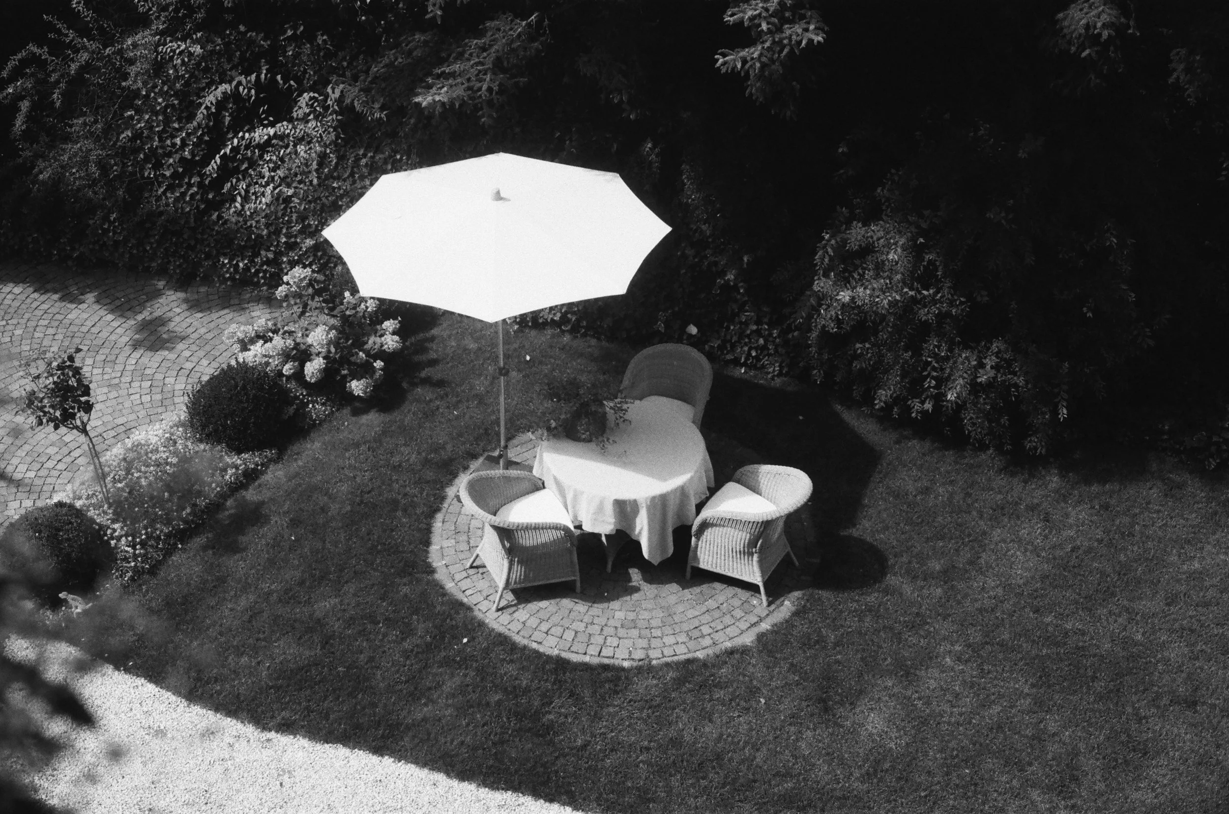 Outdoor patio area at night with a round table covered with a tablecloth, surrounded by chairs, with a large umbrella above, set on a brick-paved circular patio surrounded by grass and lush bushes.