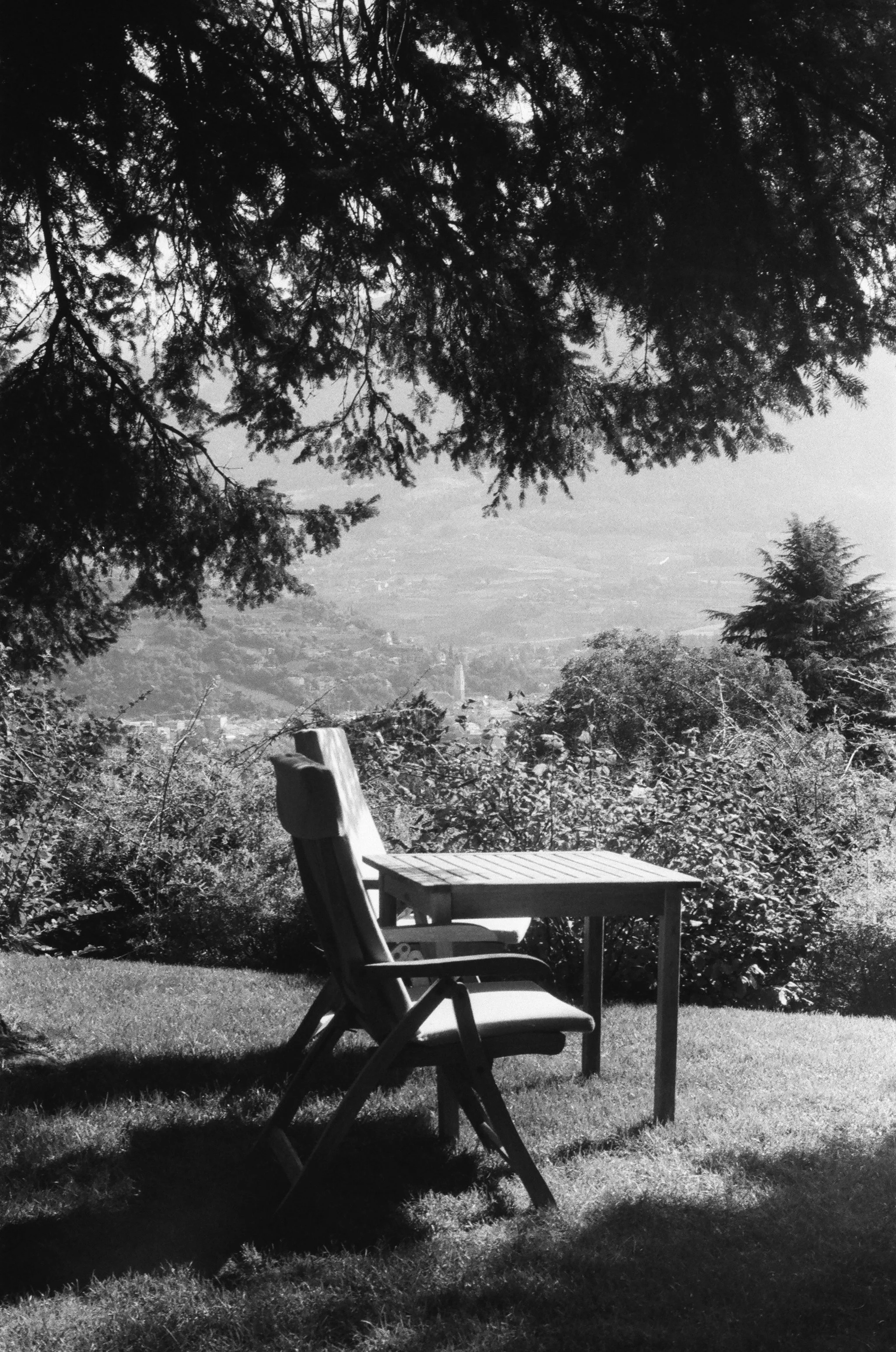 An outdoor scene with a wooden chair and table on a grassy area, trees in the background, a view of a valley and hills in the distance, in black and white.