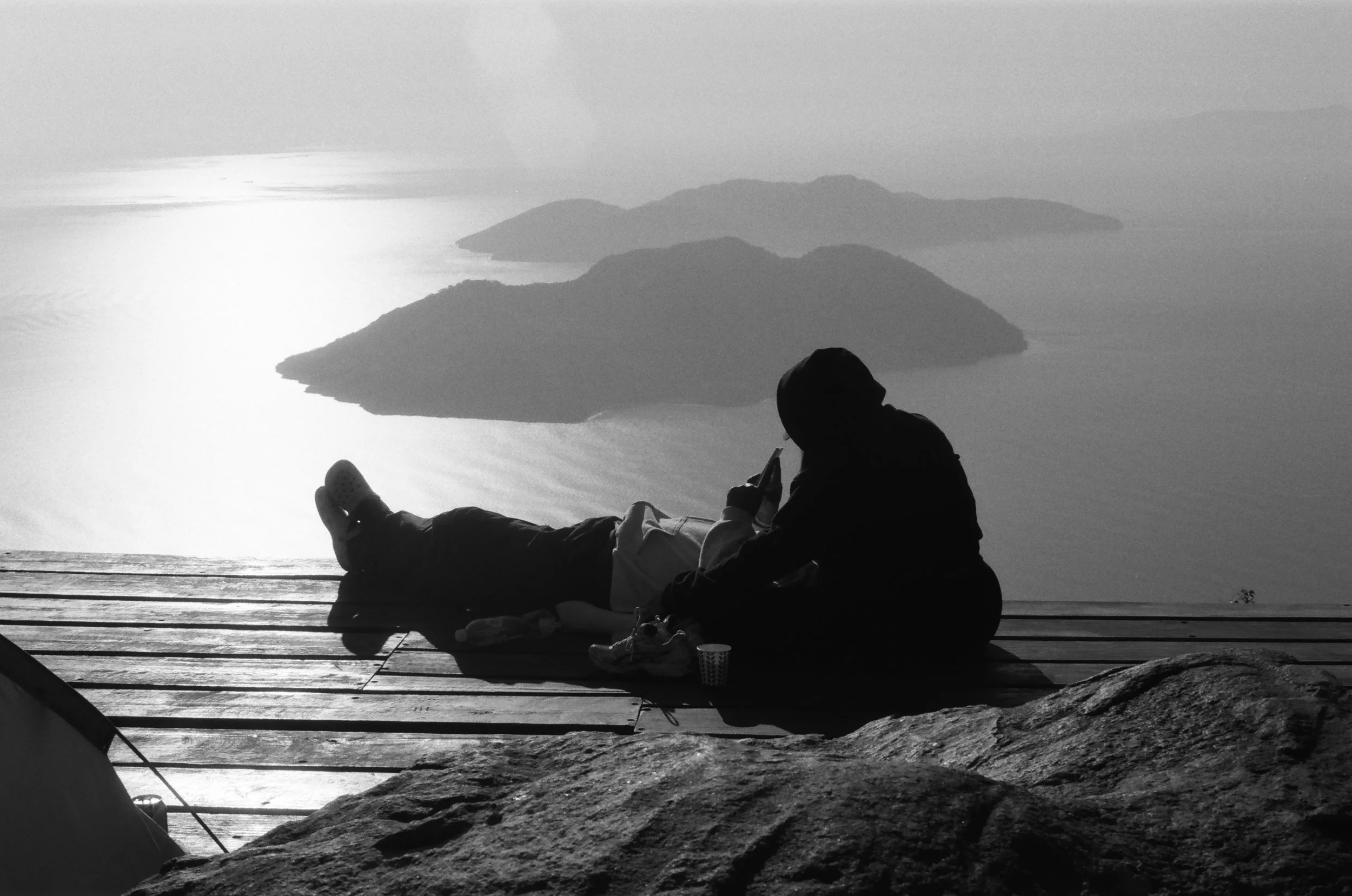 People sitting on a wooden deck overlooking a body of water and distant islands, with a person looking at their phone, in black and white.