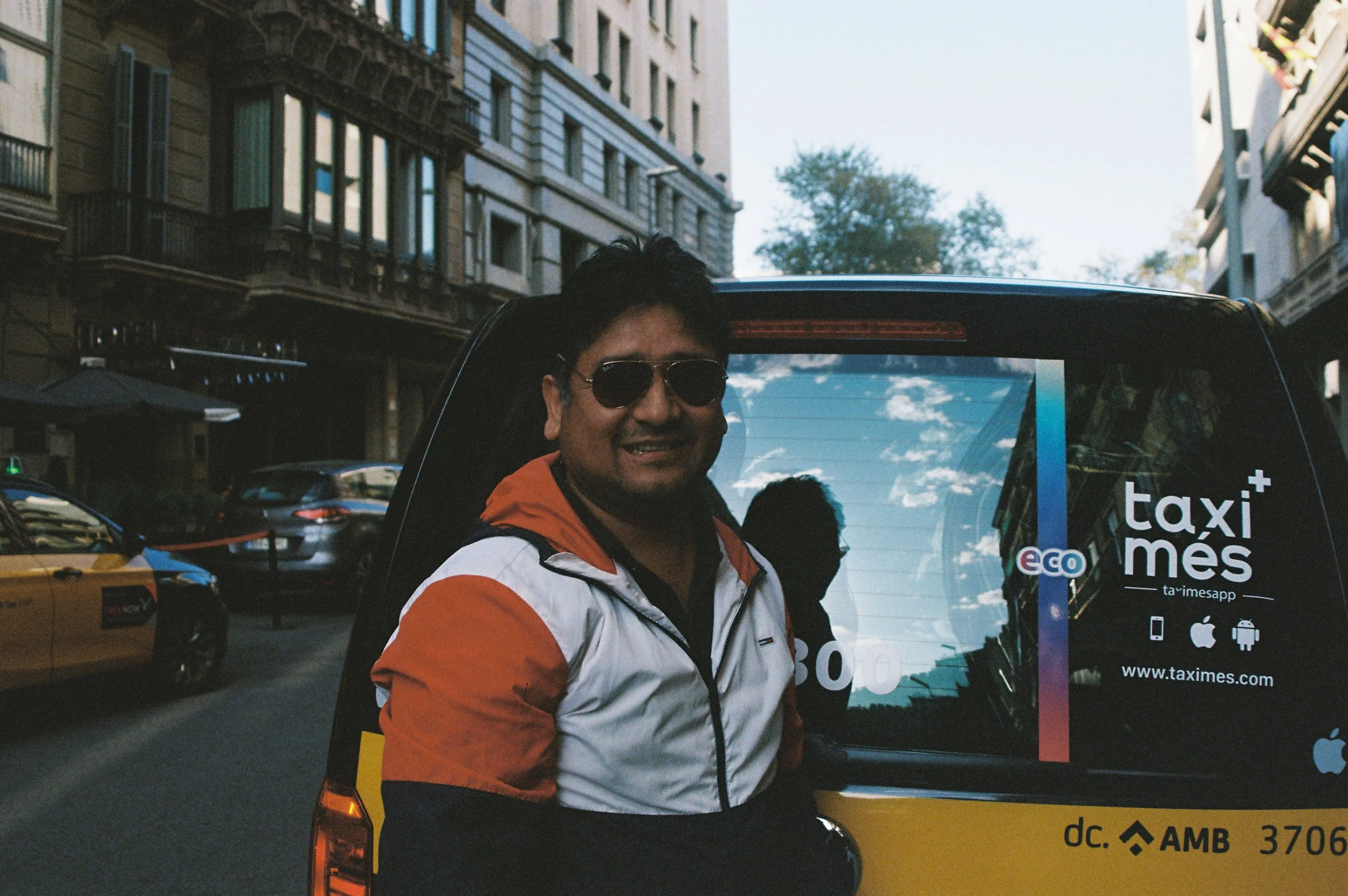 A man wearing sunglasses and an orange and white jacket standing next to a taxi with advertisements on the back window, in an urban street with tall buildings and parked cars.