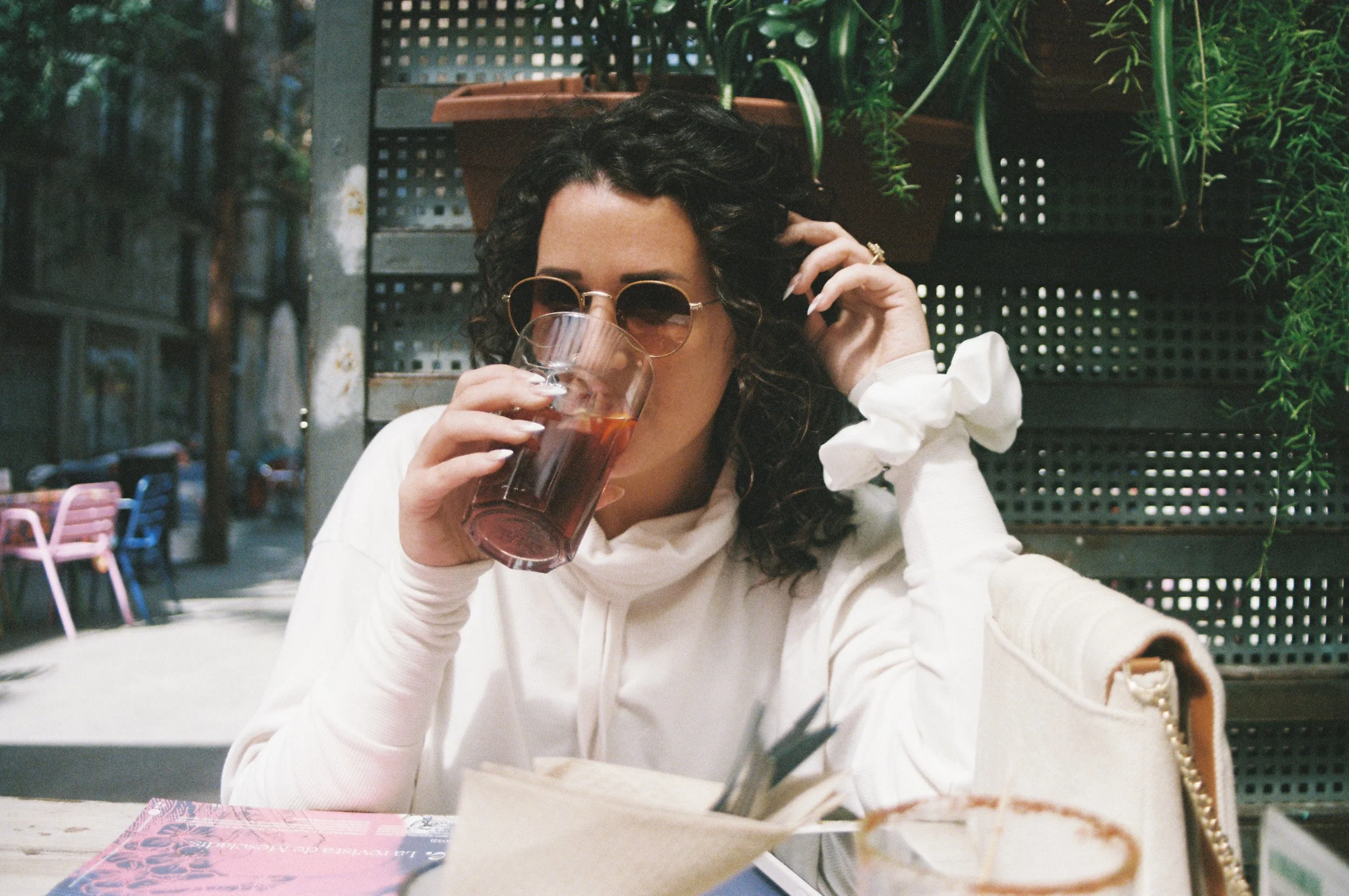 A woman with dark curly hair and sunglasses drinking iced tea at an outdoor cafe, wearing a white long-sleeve shirt, with green plants and metal chairs around her.