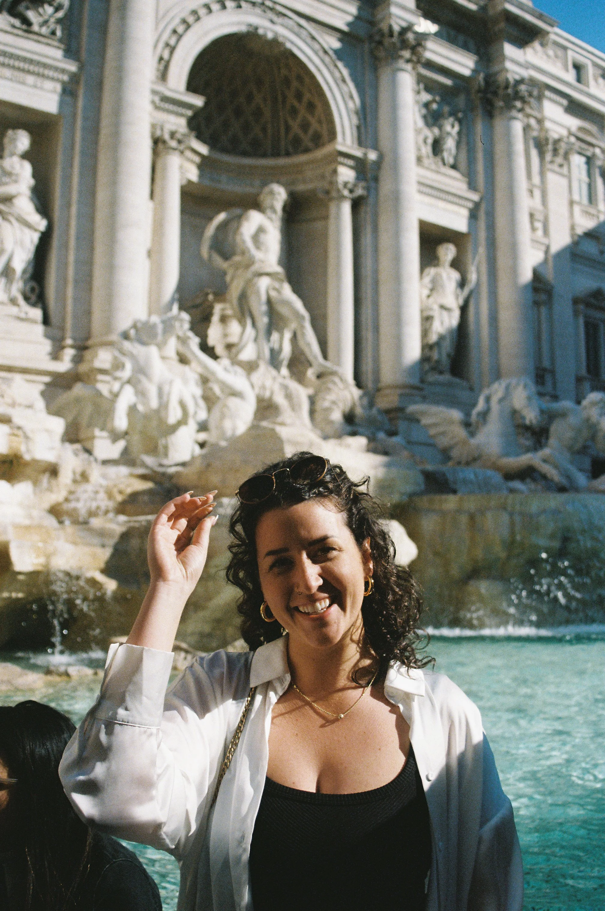 A smiling woman with dark, curly hair and gold hoop earrings, wearing a black top and white jacket, standing in front of the Trevi Fountain in Rome, Italy, with water and statues visible in the background.